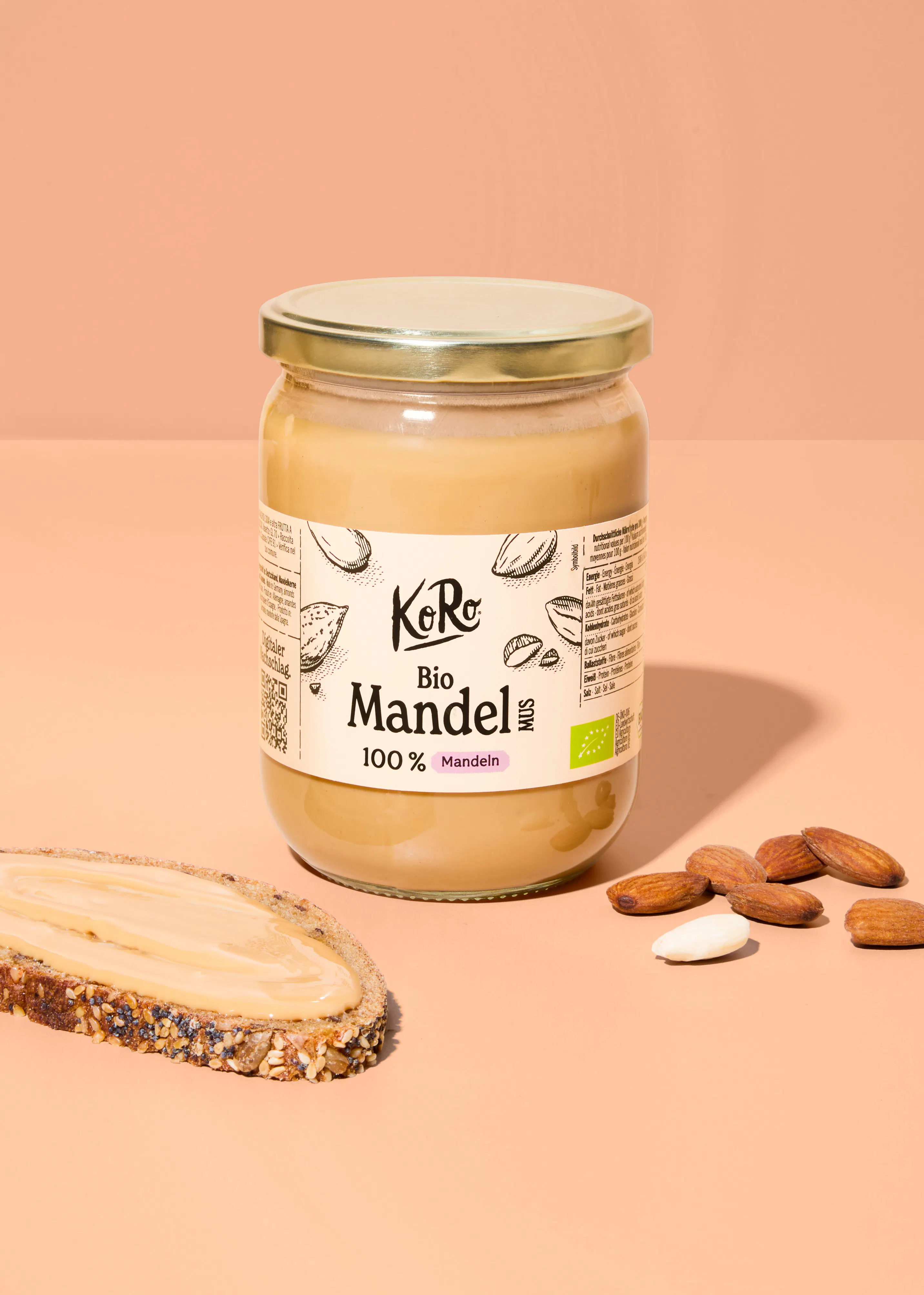 a glass jar of koro organic almond butter sits on a peach-colored surface next to a slice of seeded bread spread with the butter and a few scattered almonds.