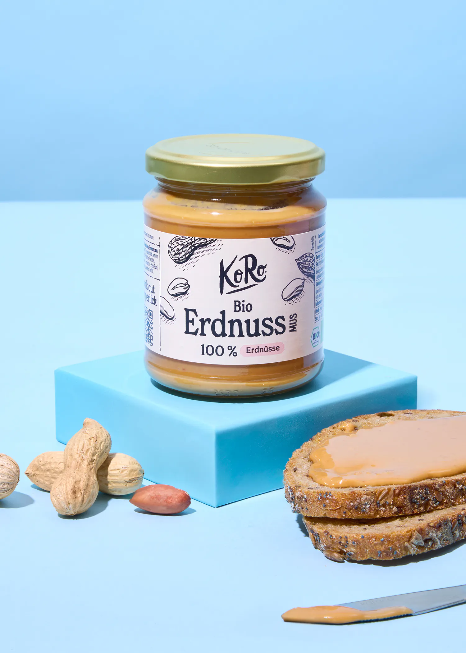 un pot de beurre de cacahuète bio koro sur une plateforme bleue, avec des cacahuètes et une tranche de pain grillé tartinée de beurre de cacahuète à côté.
