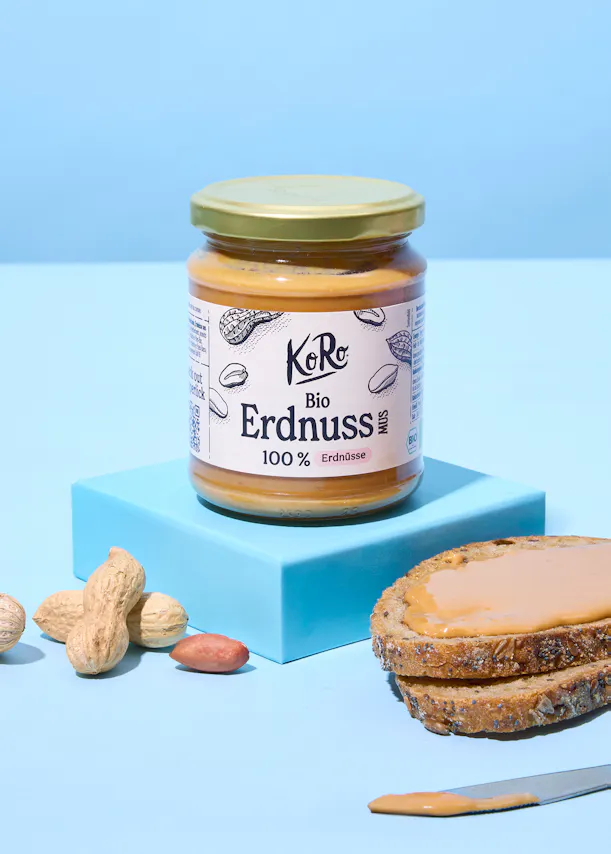 un pot de beurre de cacahuète bio koro sur une plateforme bleue, avec des cacahuètes et une tranche de pain grillé tartinée de beurre de cacahuète à côté.