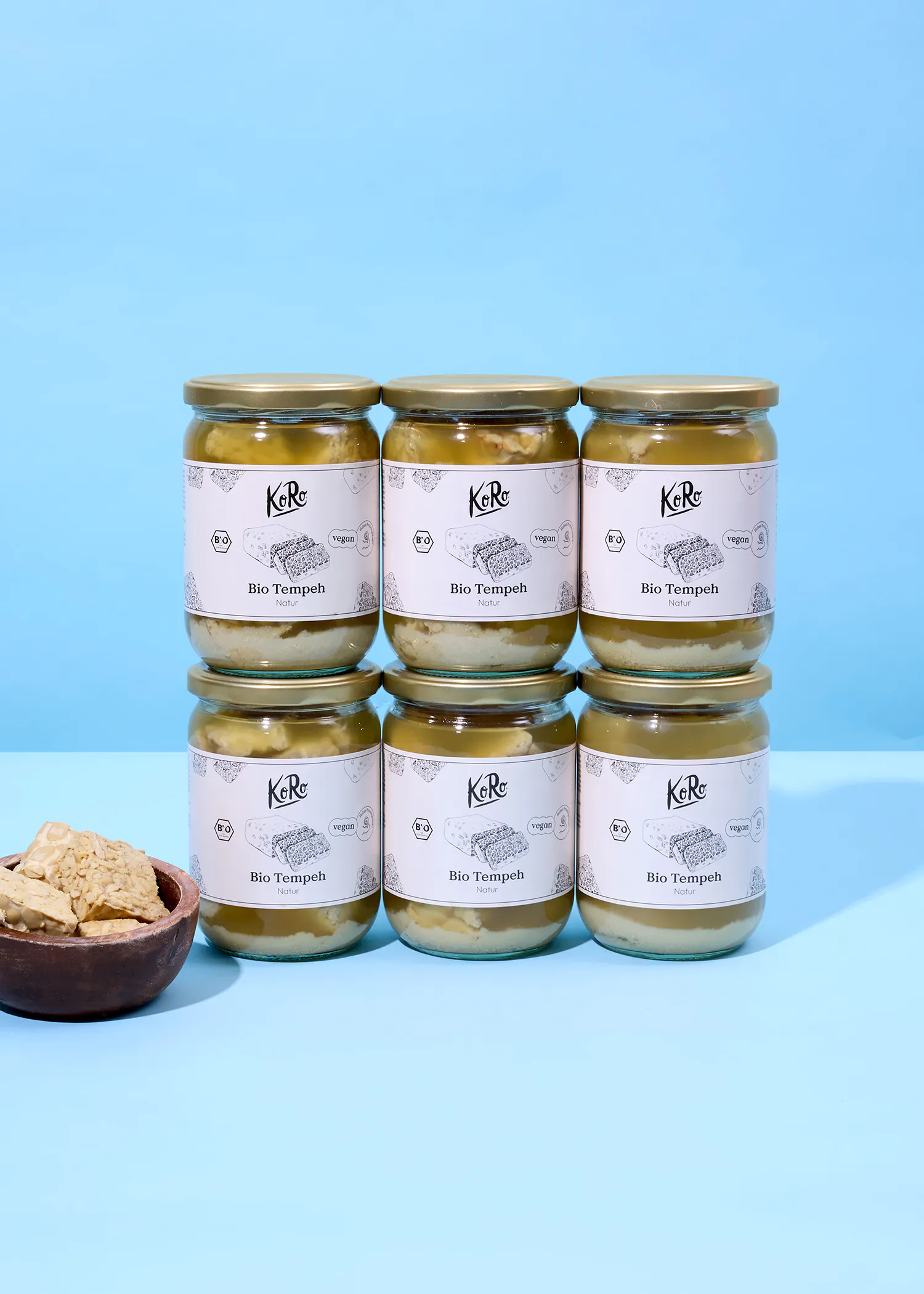 sechs gläser koro bio tempeh natur, gekennzeichnet als vegan, sind in zwei reihen gestapelt, mit einer schale tempeh-stücken im vordergrund.