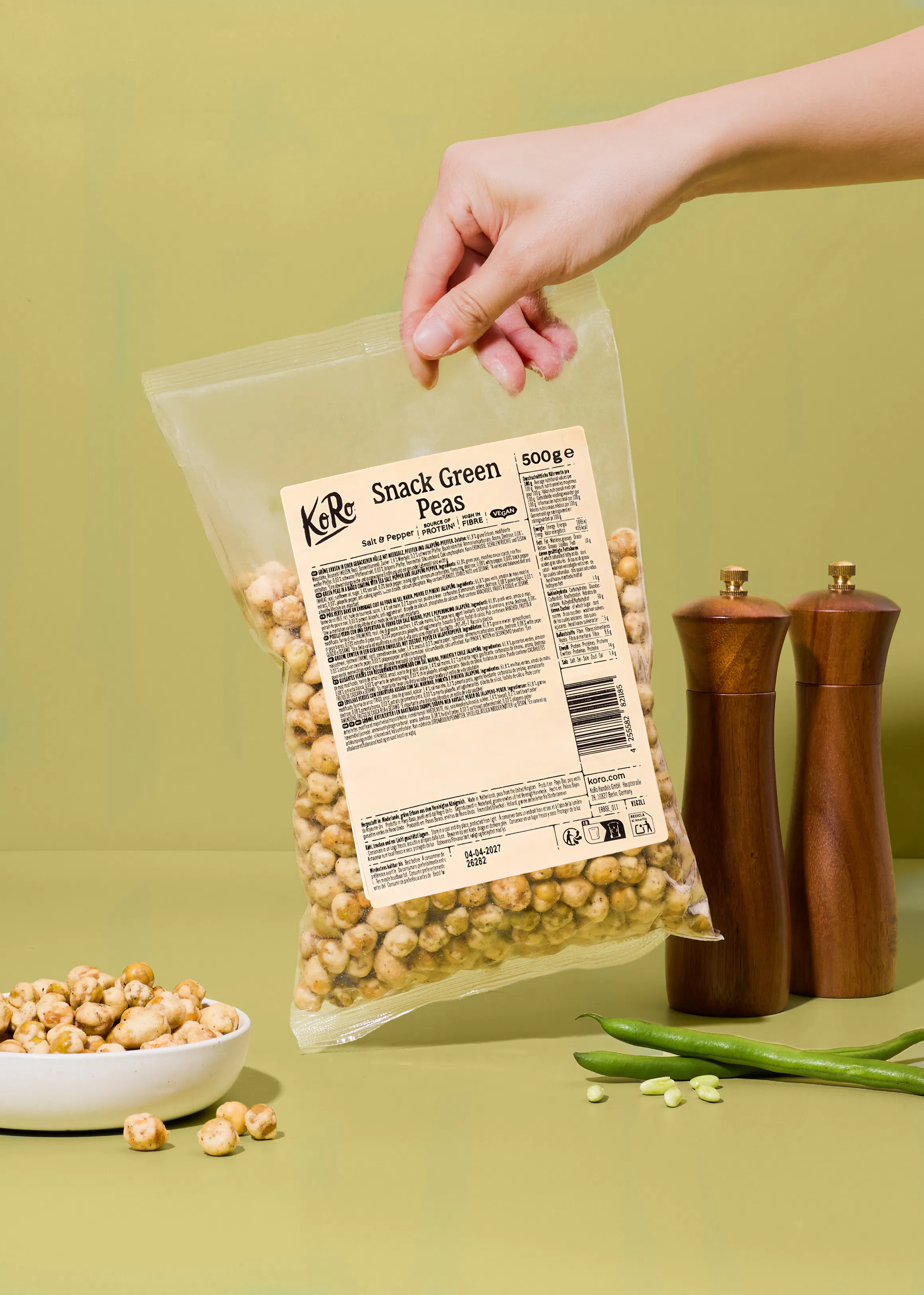 une main tient un sachet de pois verts grillés au sel et au poivre de la marque koro au-dessus d'une table avec un bol de pois et deux moulins à épices en bois.