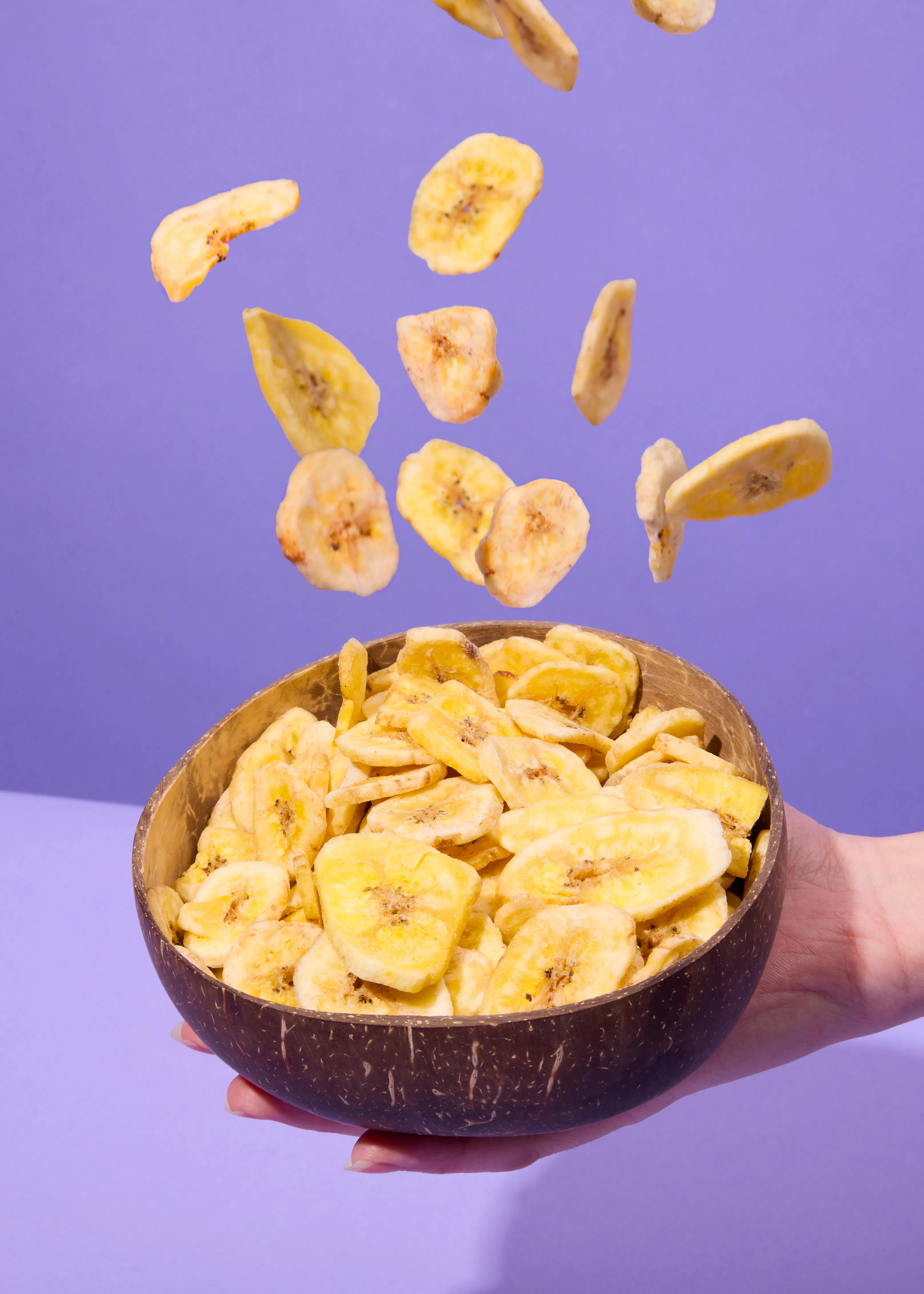 rodajas de plátano deshidratado caen en un cuenco de coco sostenido por una mano, sobre un fondo morado.