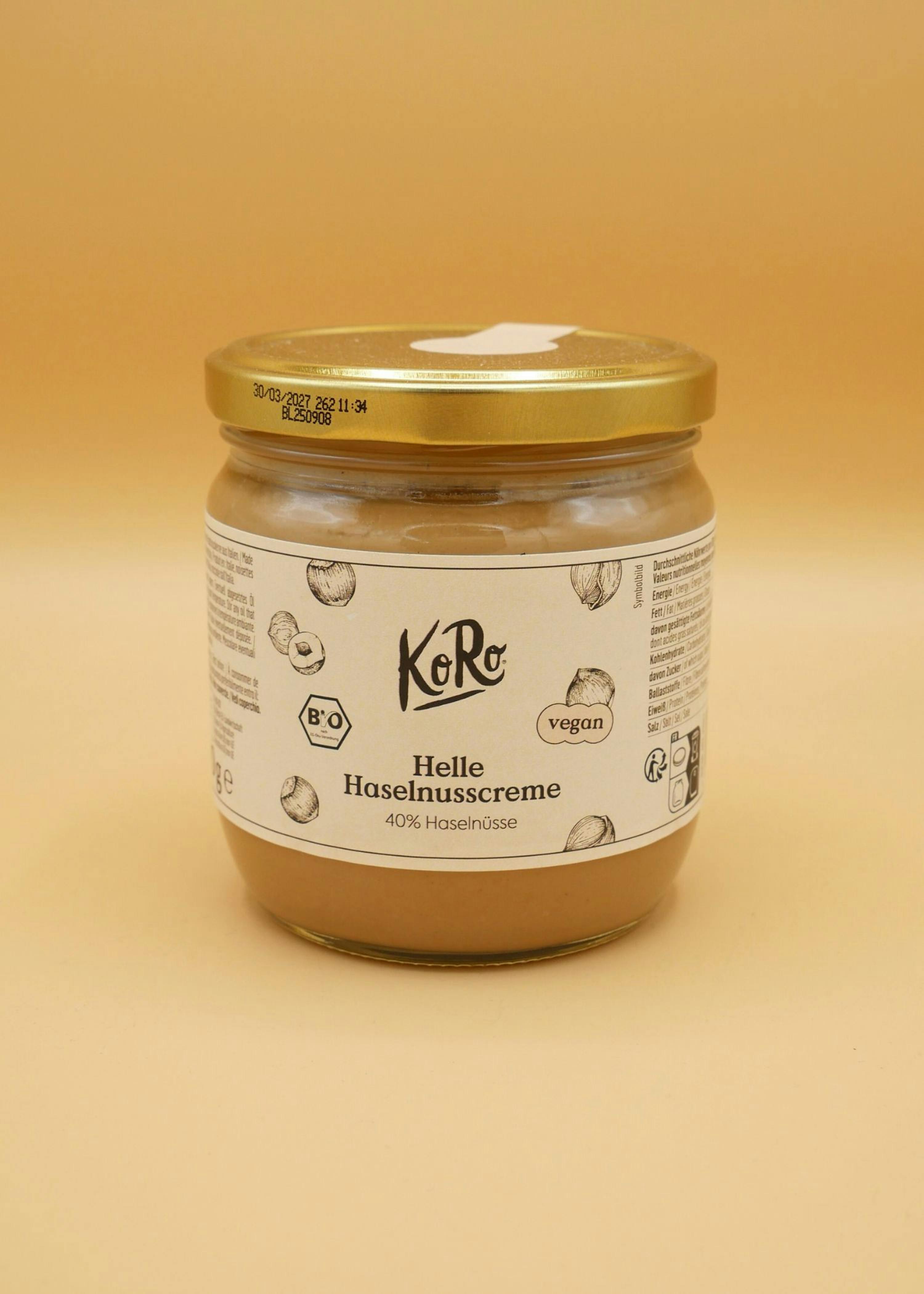 ein glas koro helle haselnusscreme mit 40% haselnüssen, bio- und vegan-zertifiziert, steht auf einem hellbraunen hintergrund.