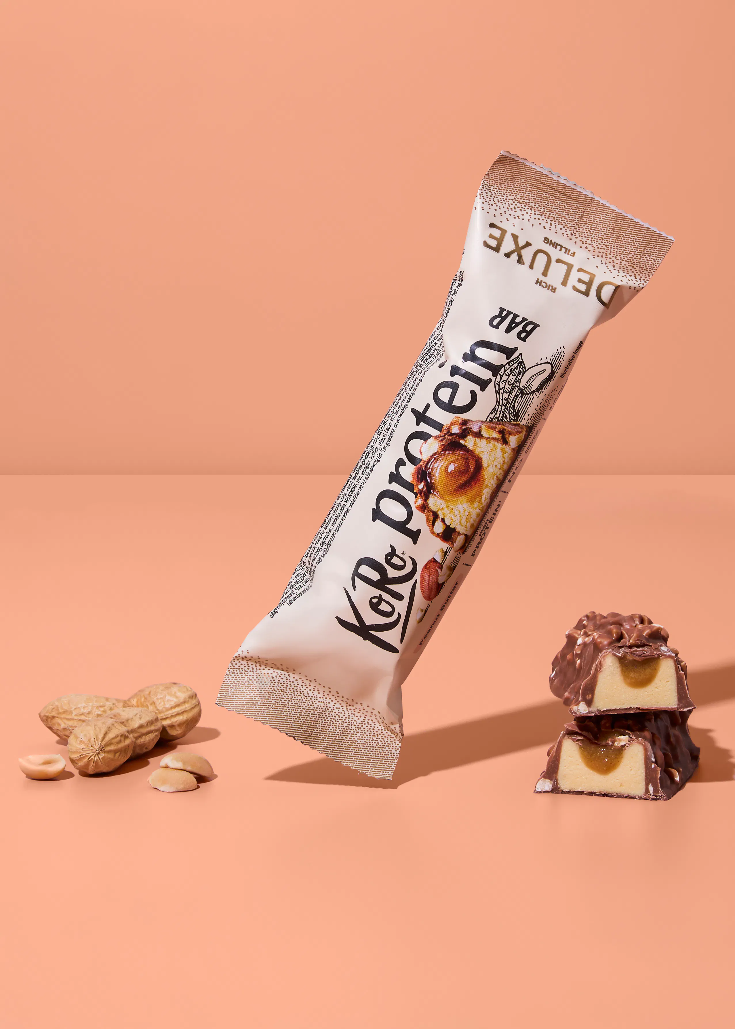 a koro protein deluxe rich filling bar in its wrapper is angled on a peach background, with whole and shelled peanuts to the left and two cut chocolate bar pieces revealing caramel and light filling to the right.