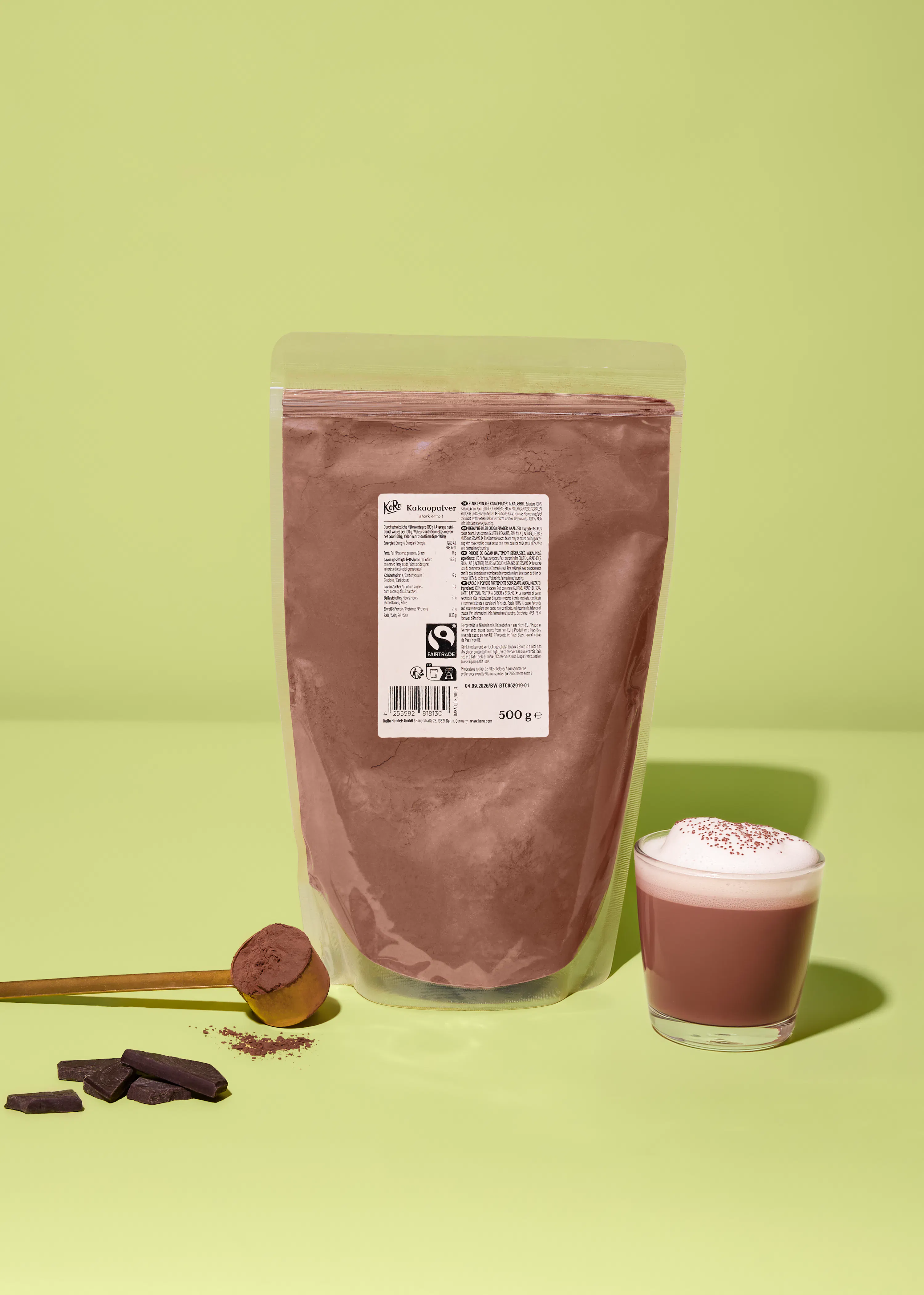 una busta di cacao in polvere da 500g con logo fairtrade, un cucchiaio di legno con cacao, pezzi di cioccolato fondente e una tazza di cioccolata calda con schiuma e granella, il tutto su uno sfondo verde chiaro.