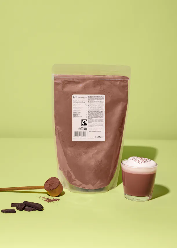 eine transparente verpackung mit kakaopulver, ein holzlöffel mit kakaopulver, schokoladenstücke und ein glas heißer kakao mit schaum auf einem hellgrünen hintergrund.