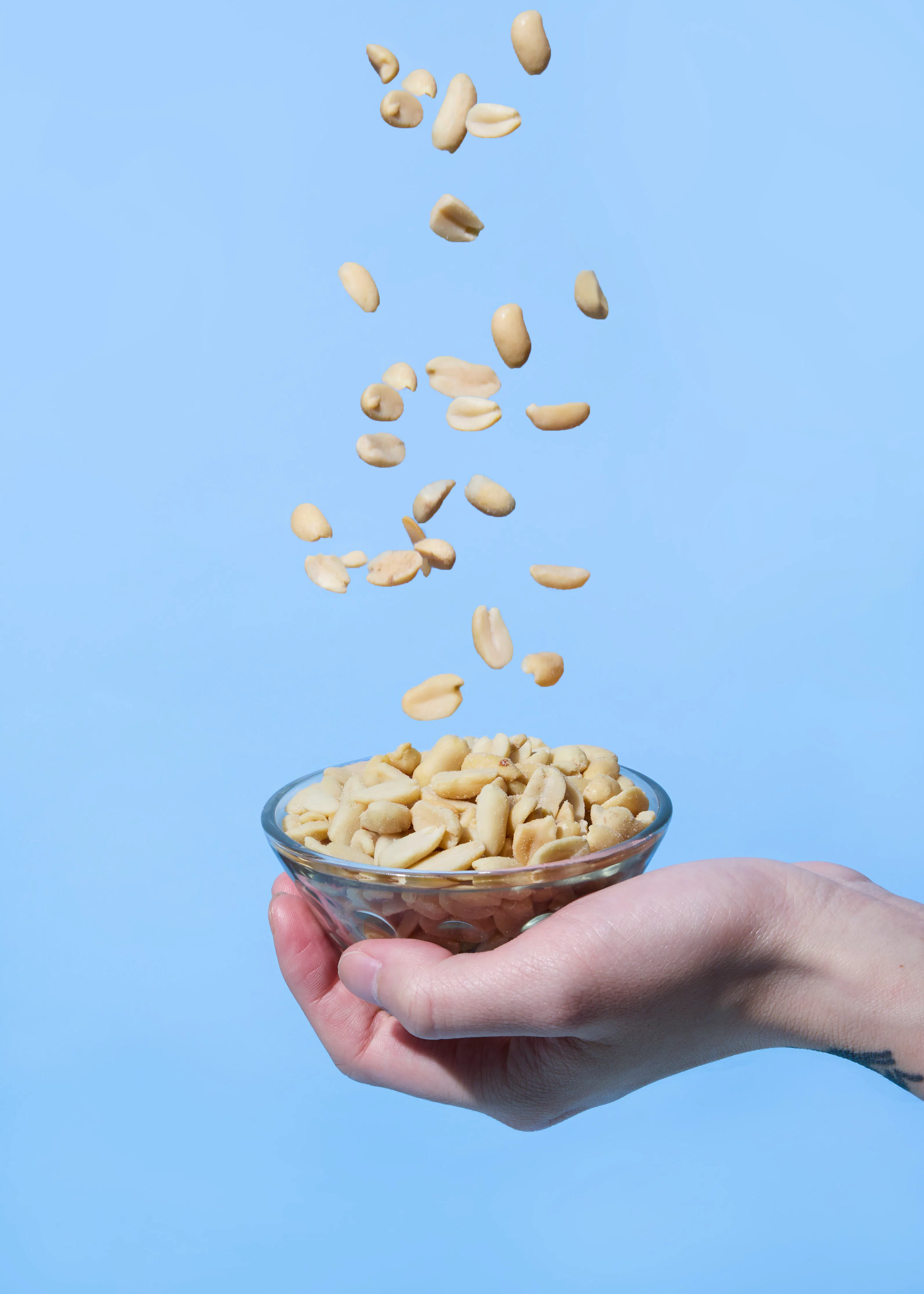une main tient un bol en verre rempli de cacahuètes, avec d'autres cacahuètes tombant dans le bol sur un fond bleu uni.
