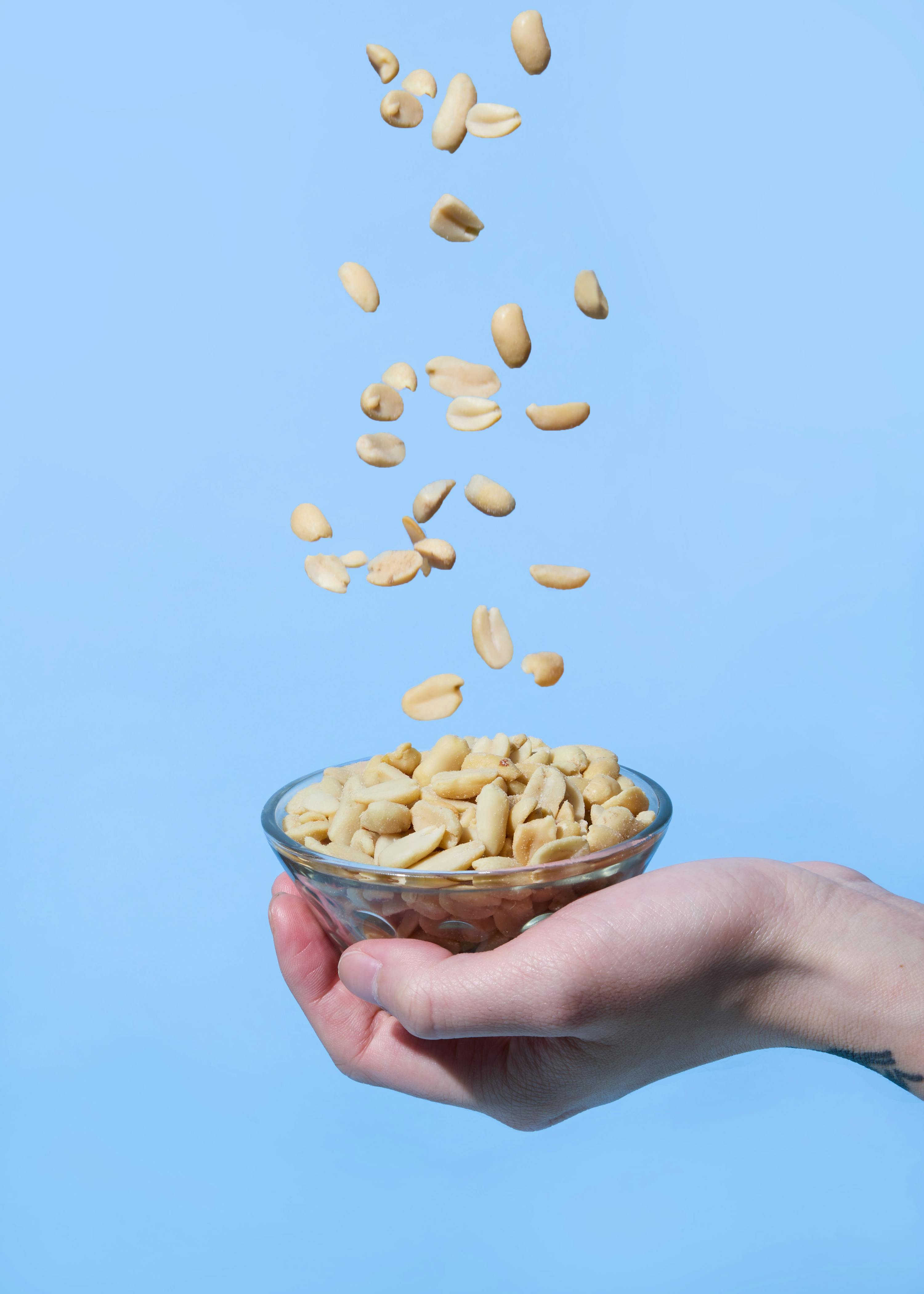 erdnüsse fallen in eine von einer hand gehaltene glasschale vor blauem hintergrund.