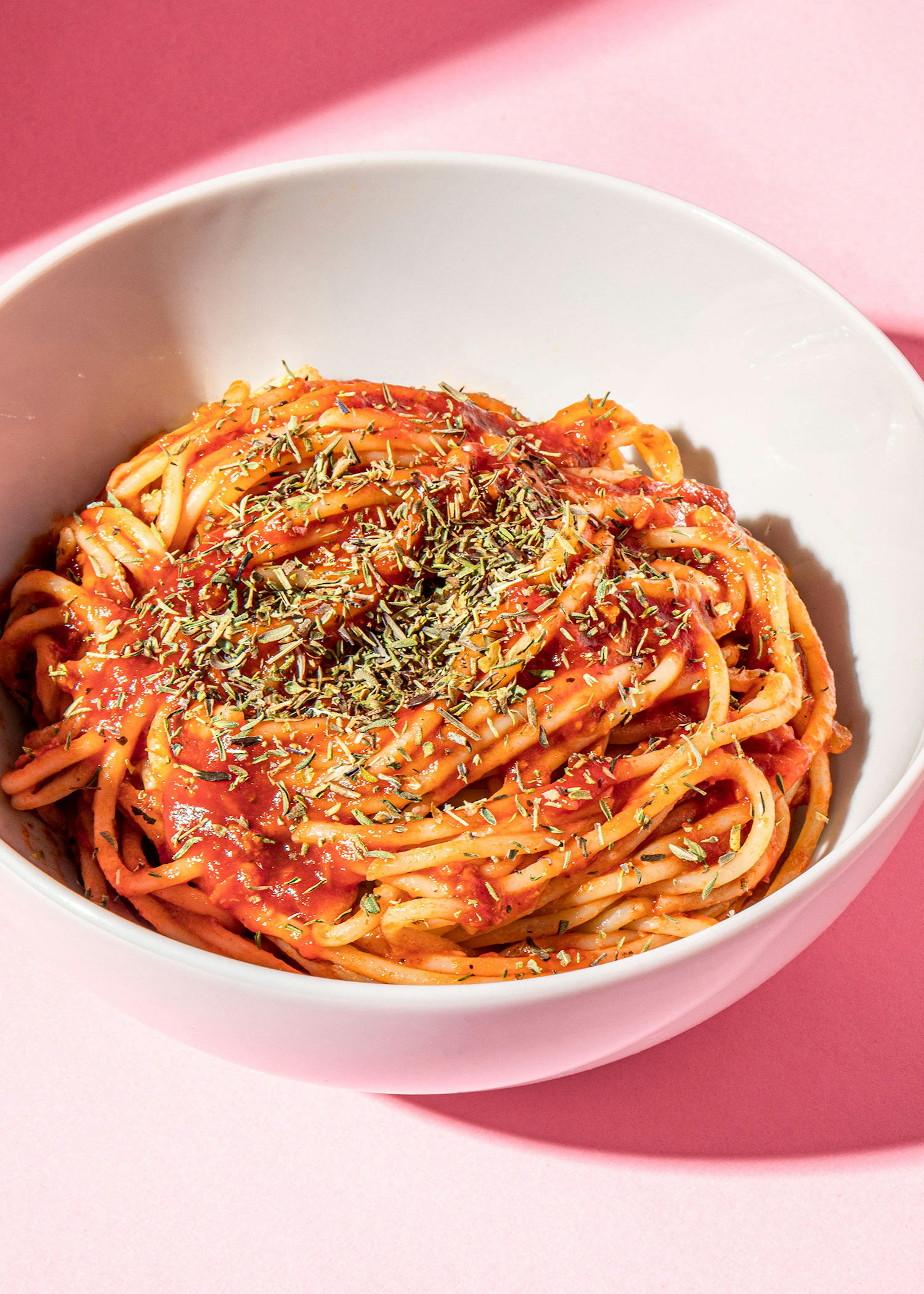 eine weiße Schüssel Nudeln mit Tomatensauce und Kräutern auf einem rosa Hintergrund
