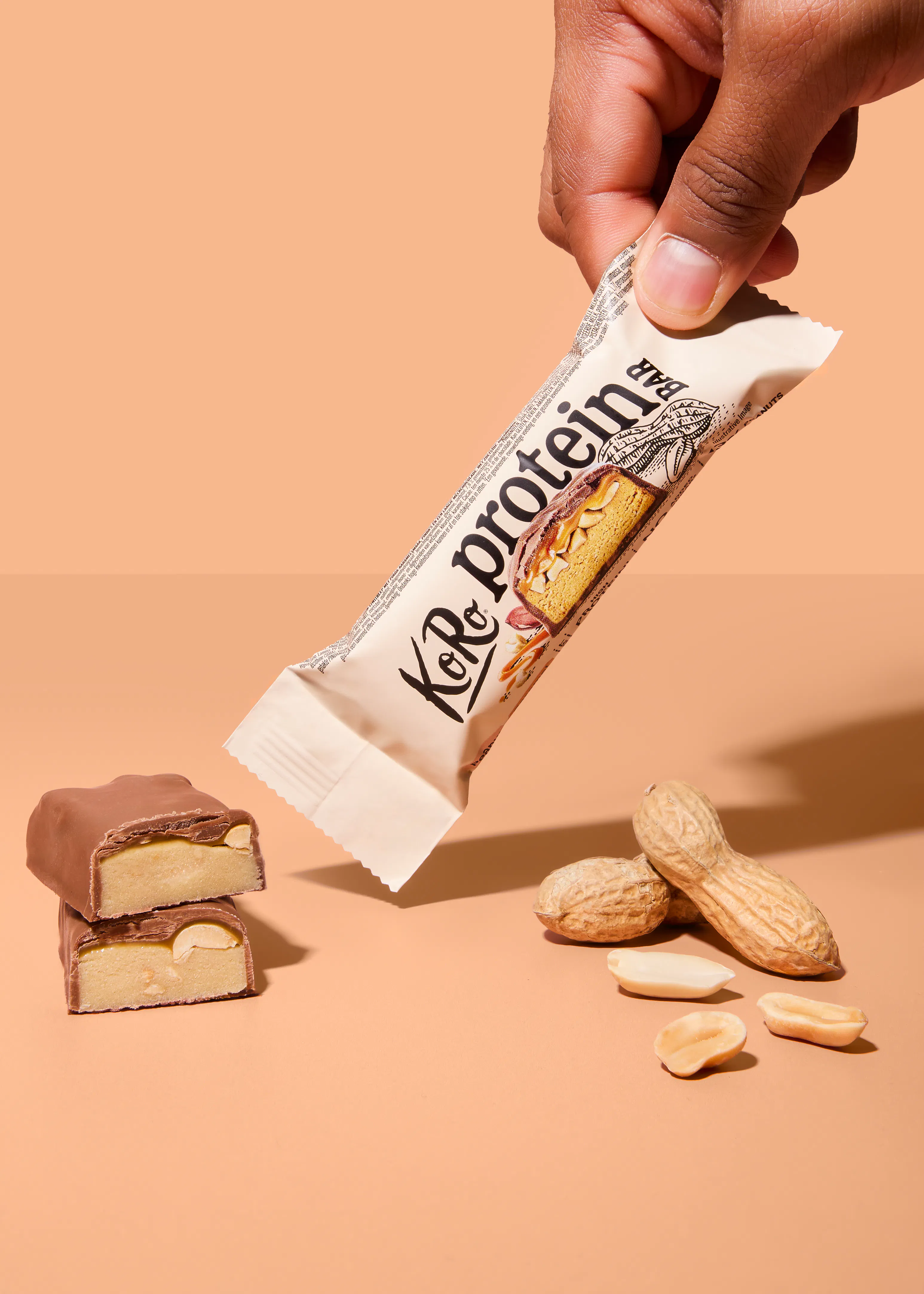une main tient un emballage de barre protéinée koro avec une illustration de la barre coupée, tandis que deux moitiés de barre chocolatée et des cacahuètes entières et décortiquées sont posées sur une surface beige.