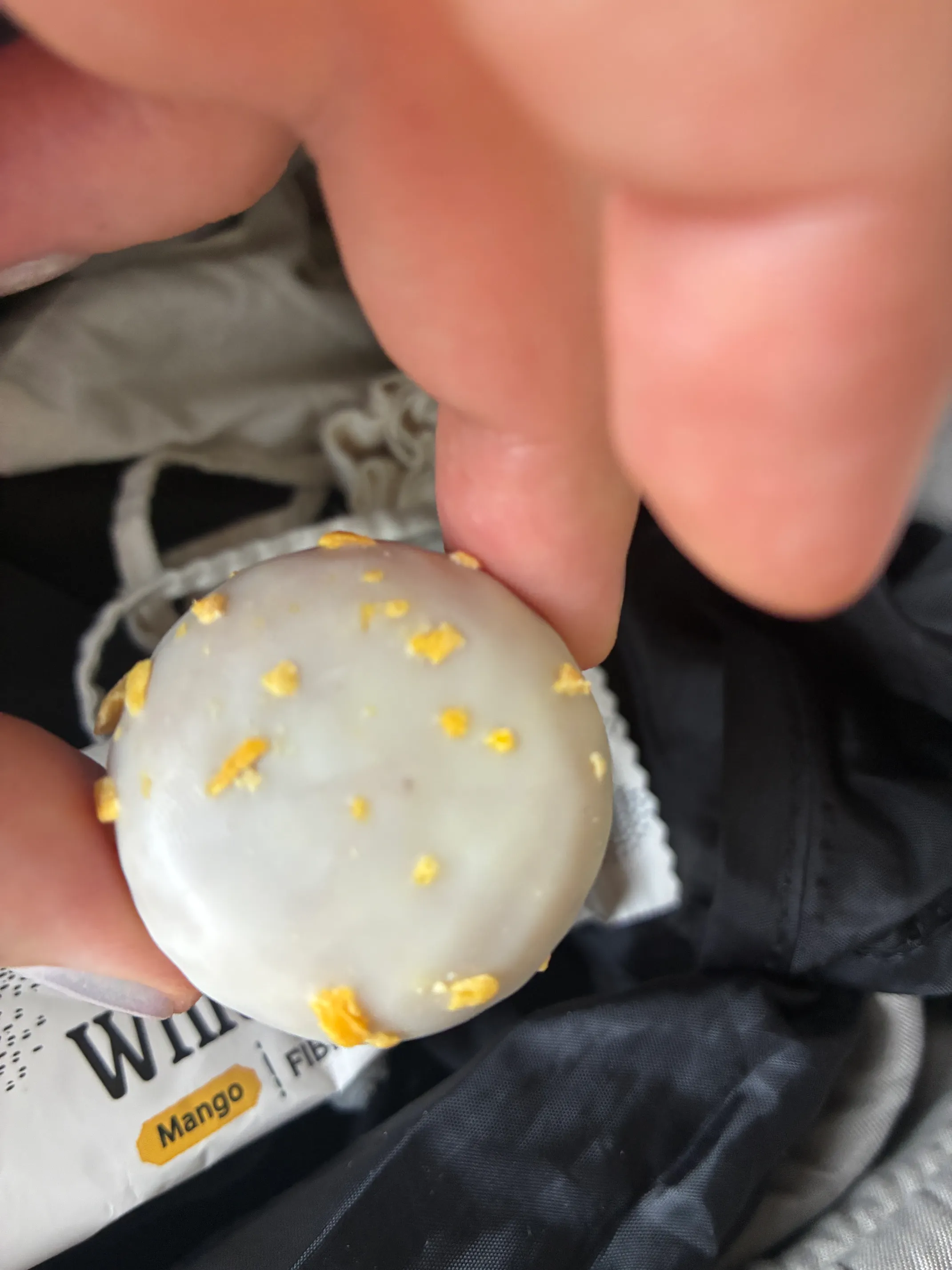 una mano tiene uno snack rotondo glassato di bianco con pezzetti gialli, con uno sfondo sfocato che include una confezione con la scritta "mango".