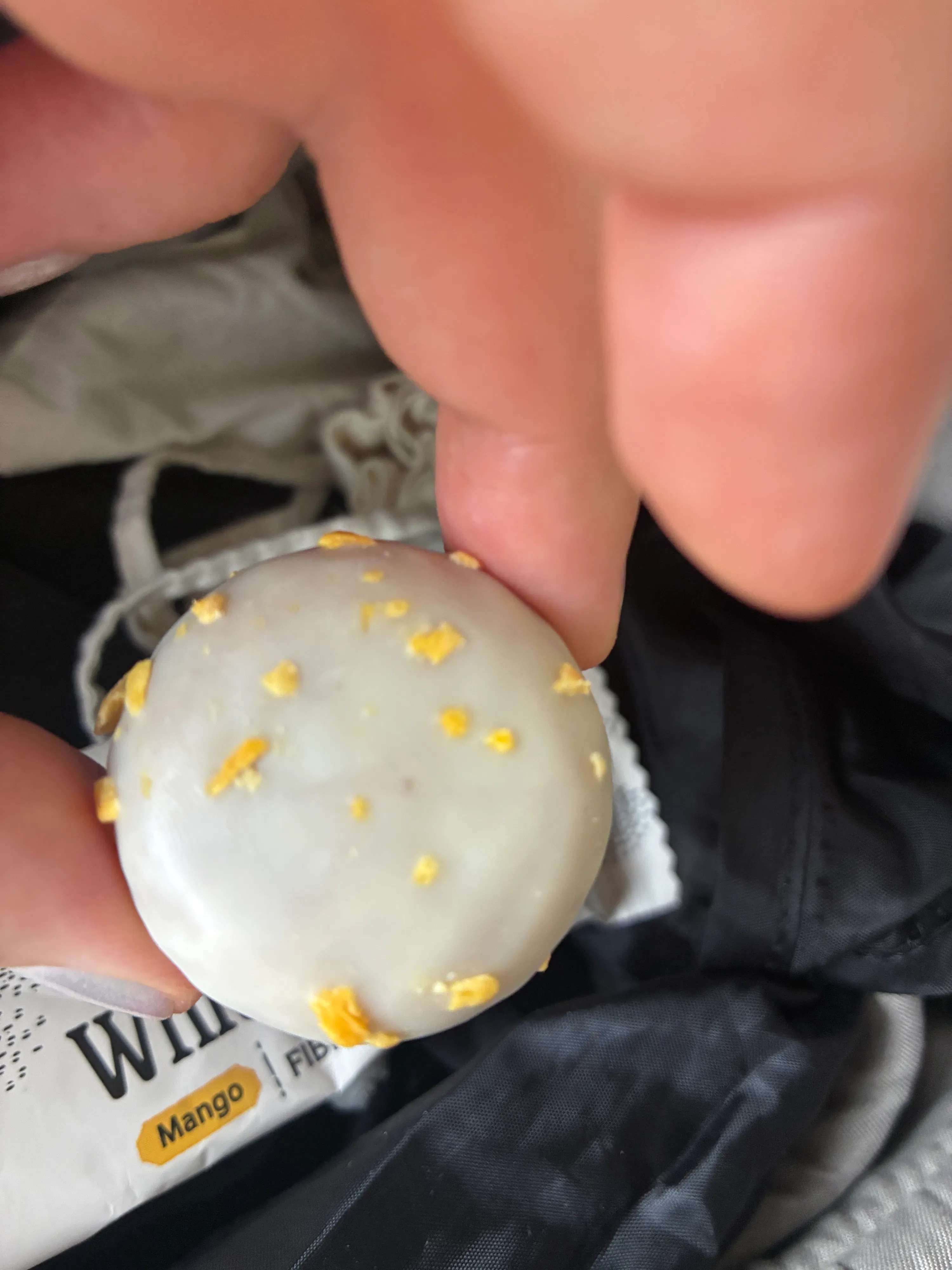 une main tient une collation ronde glacée de blanc avec des éclats jaunes, avec un emballage de mangue en arrière-plan.