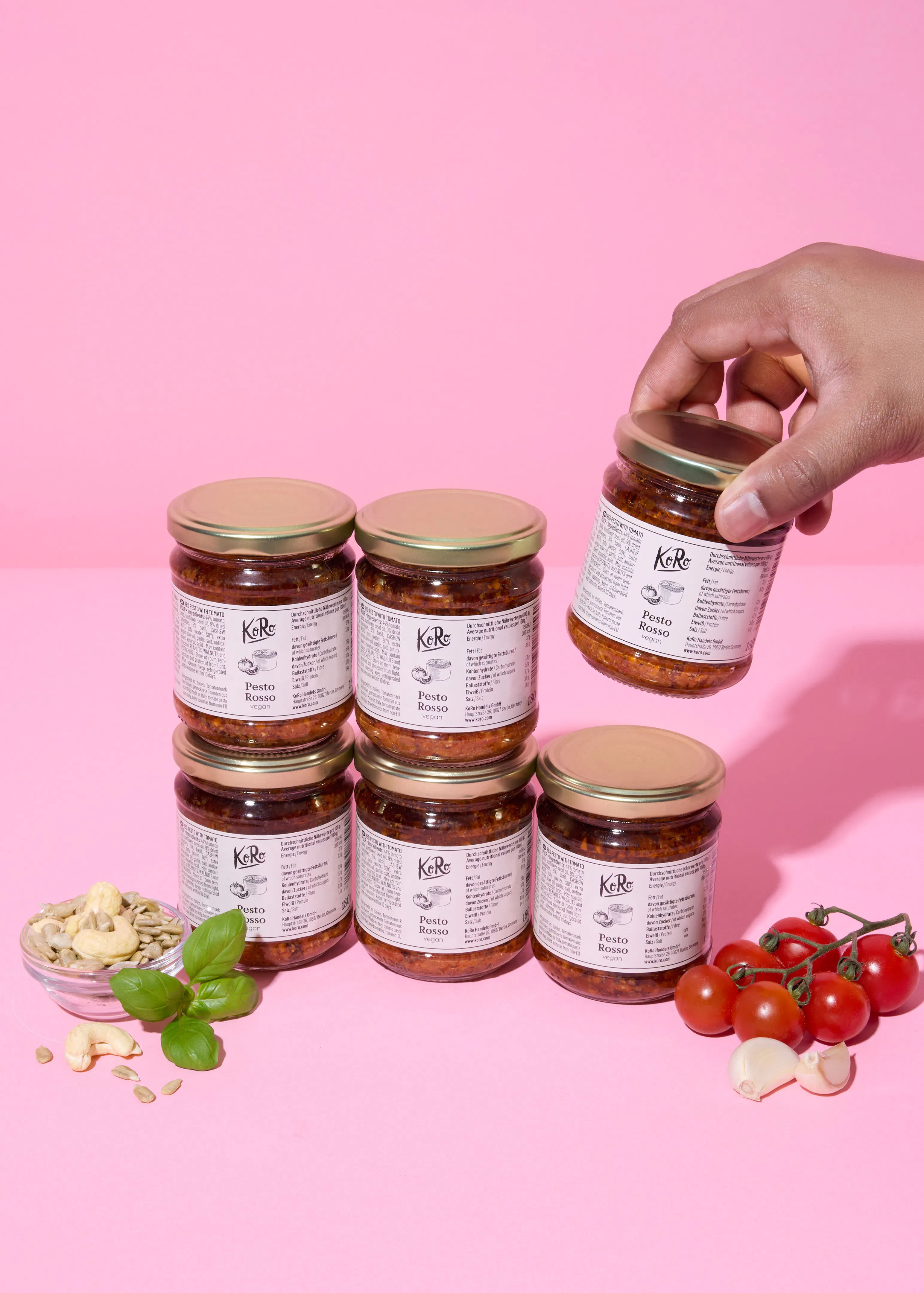 a hand holds a jar of pesto rosso above several other jars of pesto rosso, with cherry tomatoes, garlic, basil, cashews, and sunflower seeds on a pink background.