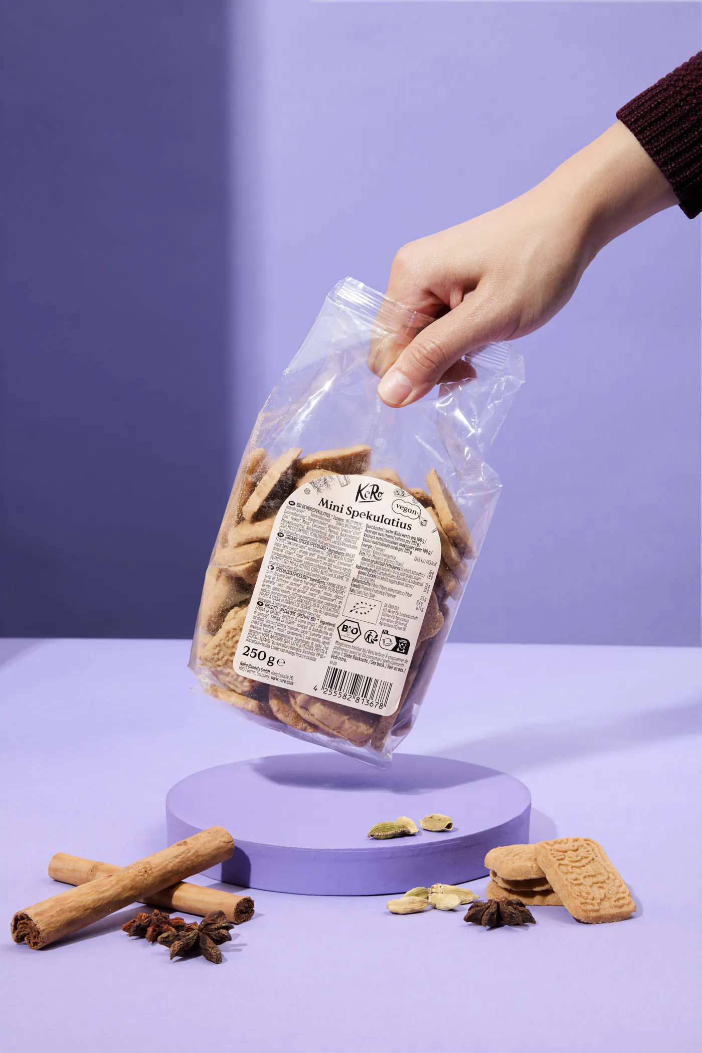 a hand holds a clear bag of mini spekulatius cookies over a purple surface with cinnamon sticks, star anise, and cardamom pods.