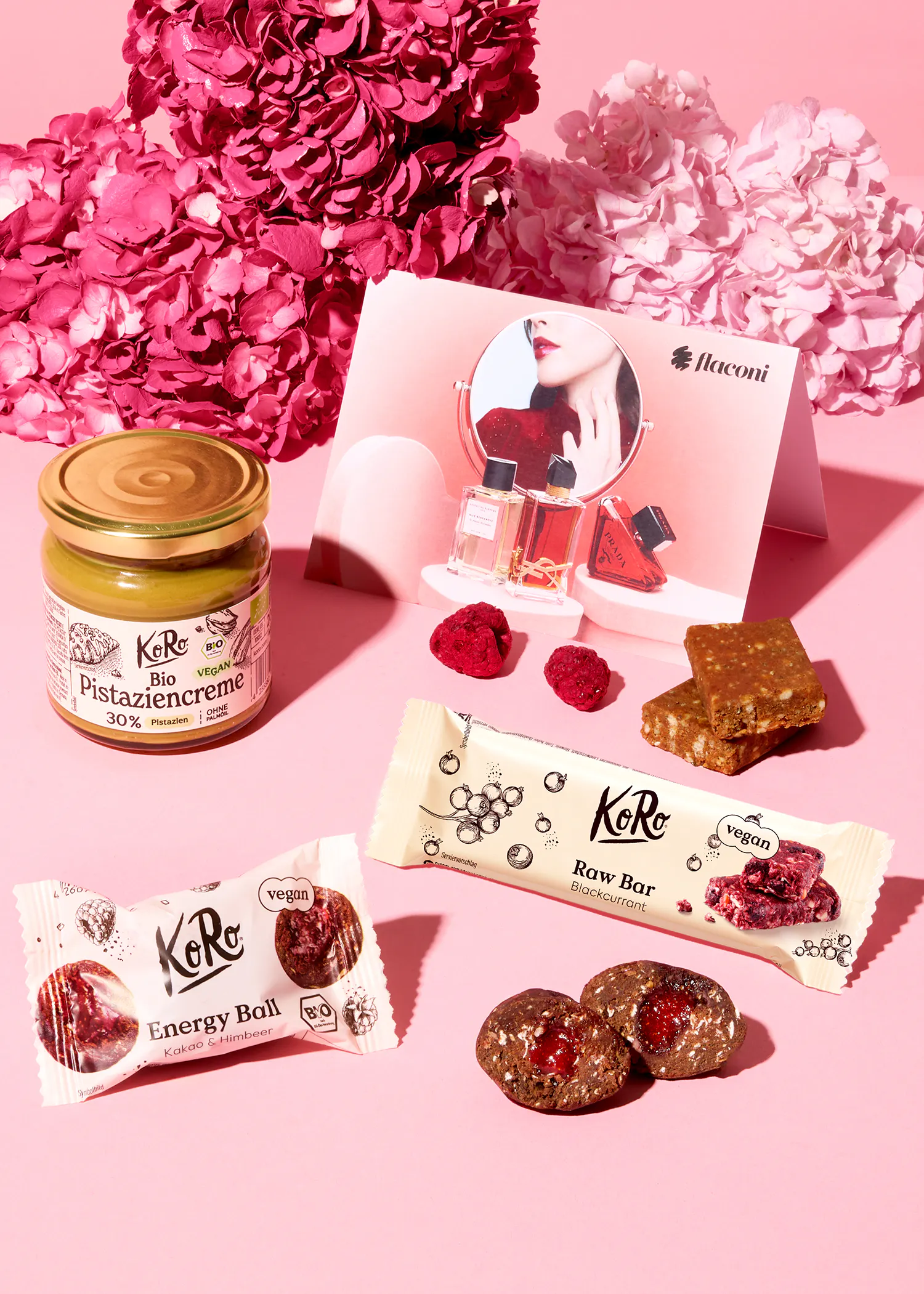 ein stillleben mit koro-produkten wie pistaziencreme, energiekugeln und riegeln, getrockneten himbeeren, einer flaconi-werbekarte und rosa blumen auf einem pinken hintergrund.