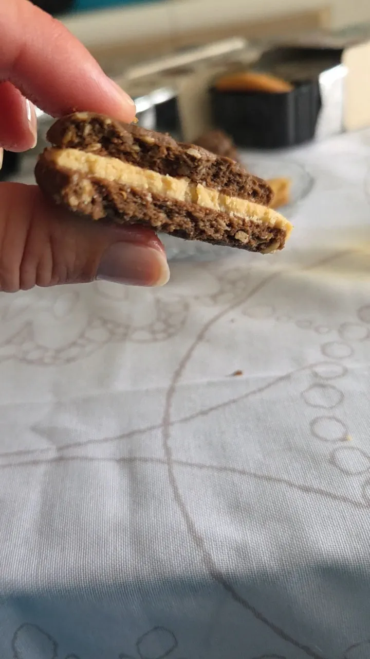 una mano tiene un biscotto al cioccolato farcito con una crema chiara su una tovaglia bianca.
