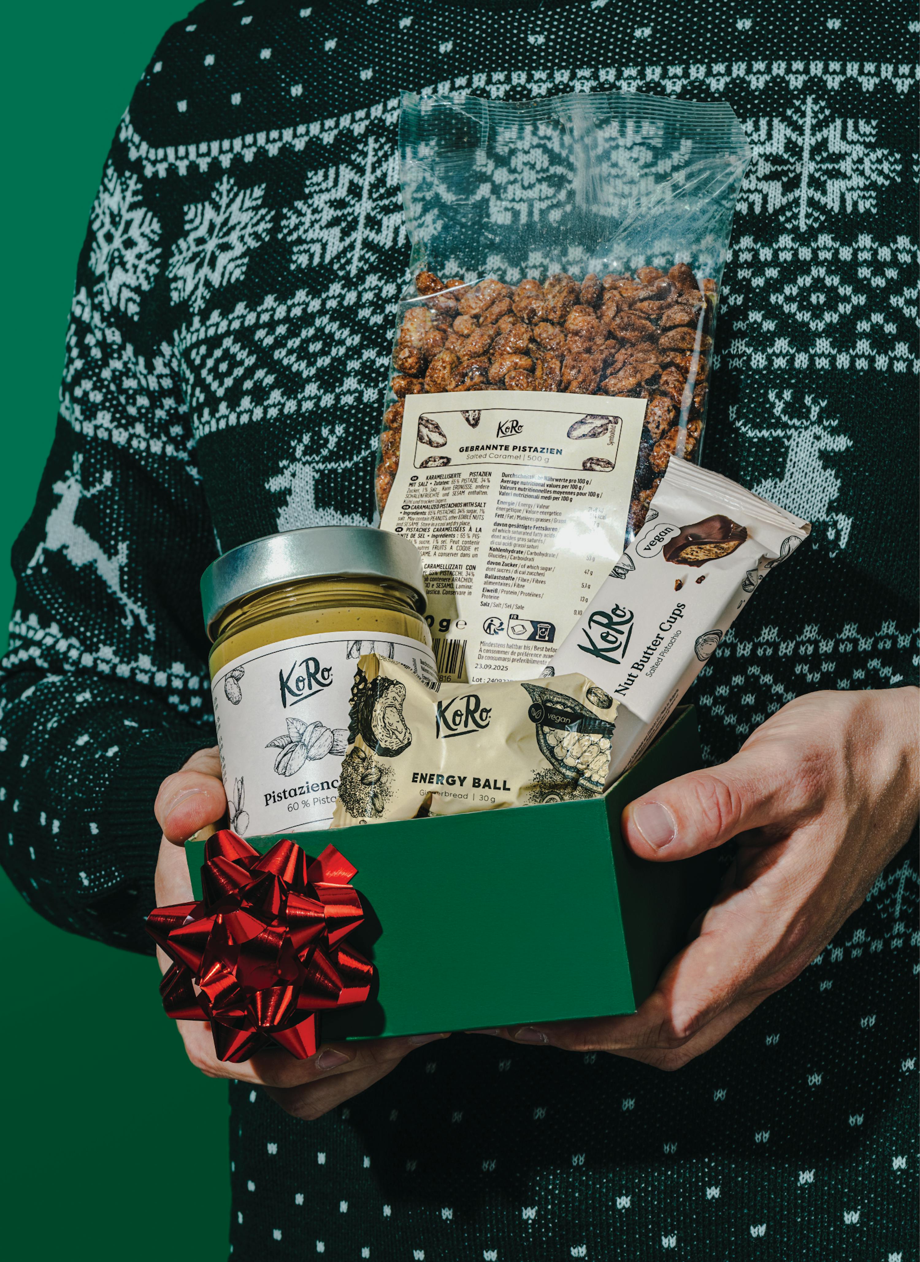 une personne portant un pull de noël tient une boîte cadeau verte ornée d'un ruban rouge, remplie de produits ko ro à la pistache, dont des pistaches caramélisées, une pâte à tartiner, une boule d'énergie et une barre de beurre de noix.