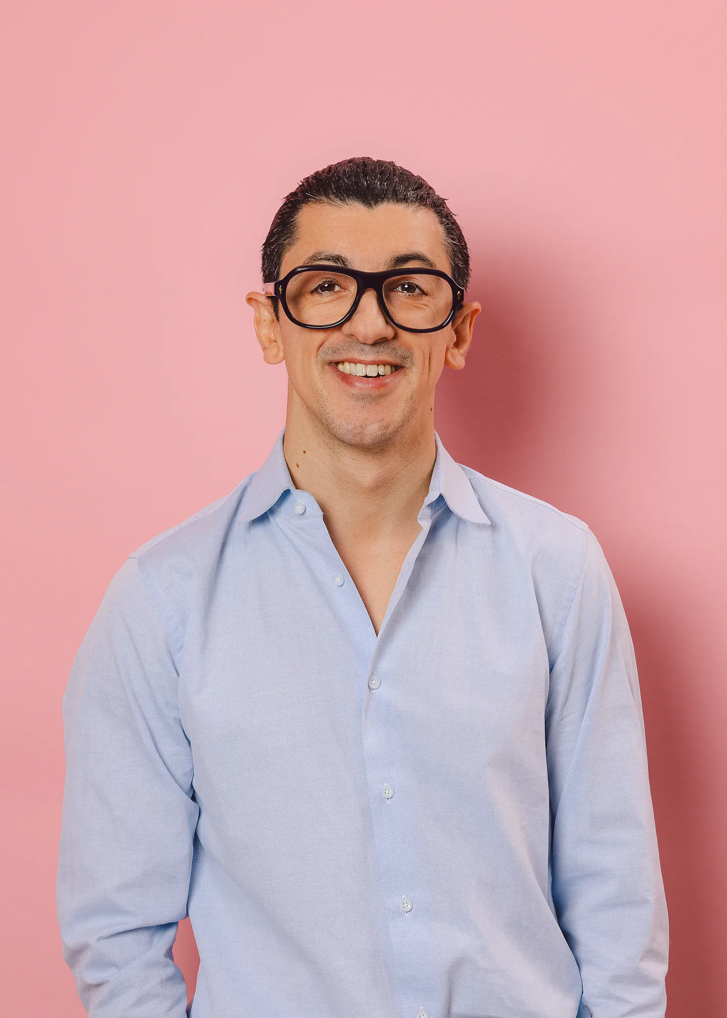 un uomo con gli occhiali e la camicia blu sorride davanti a uno sfondo rosa