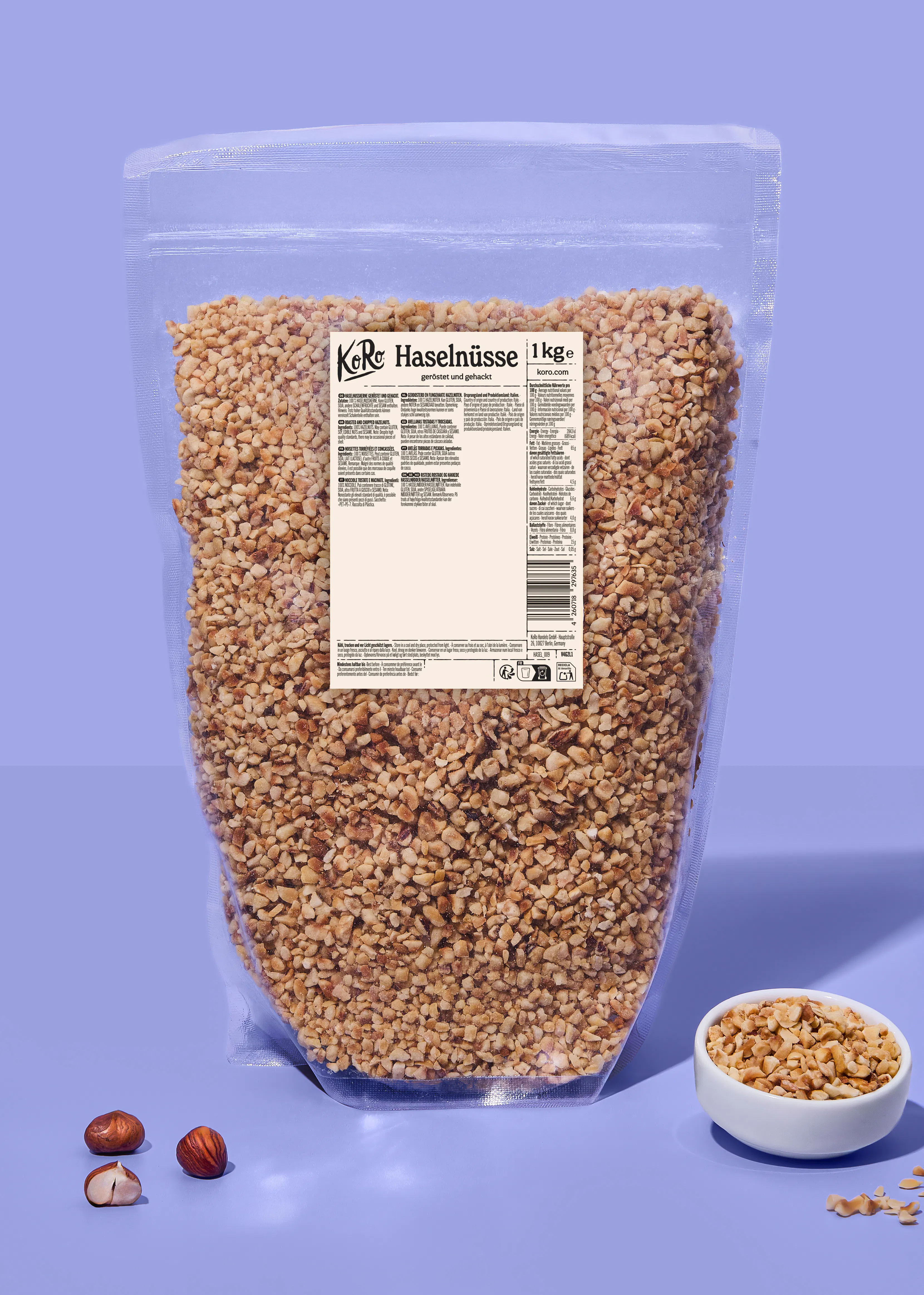 a one-kilogram clear plastic bag of roasted and chopped hazelnuts from koro, with a small bowl of the nuts and two whole hazelnuts placed in front on a light purple background.