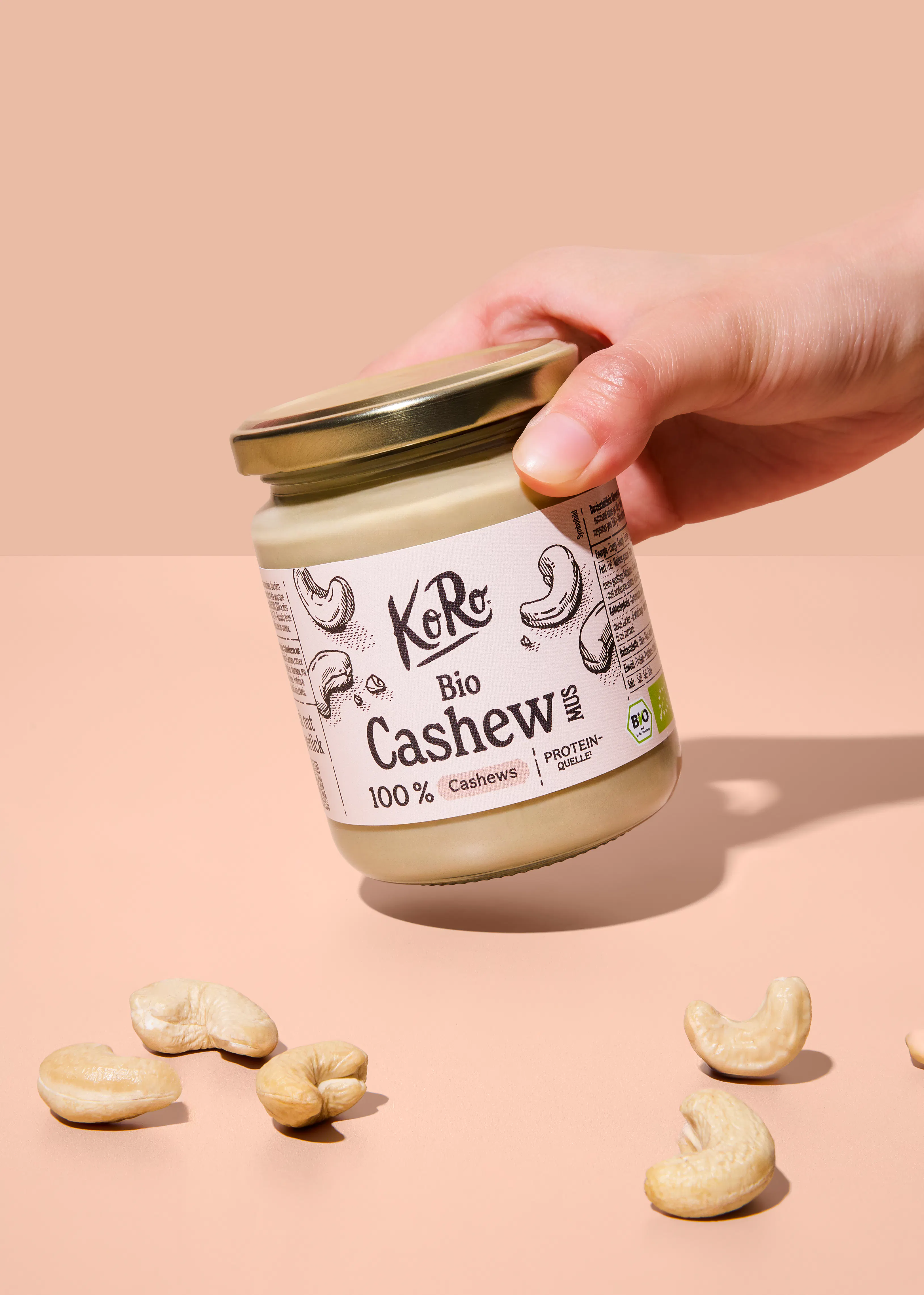 eine hand hält ein glas koro bio cashew mus, beschriftet mit "100% cashews" und "protein-quelle", während ganze cashewnüsse auf einer hellen oberfläche liegen.