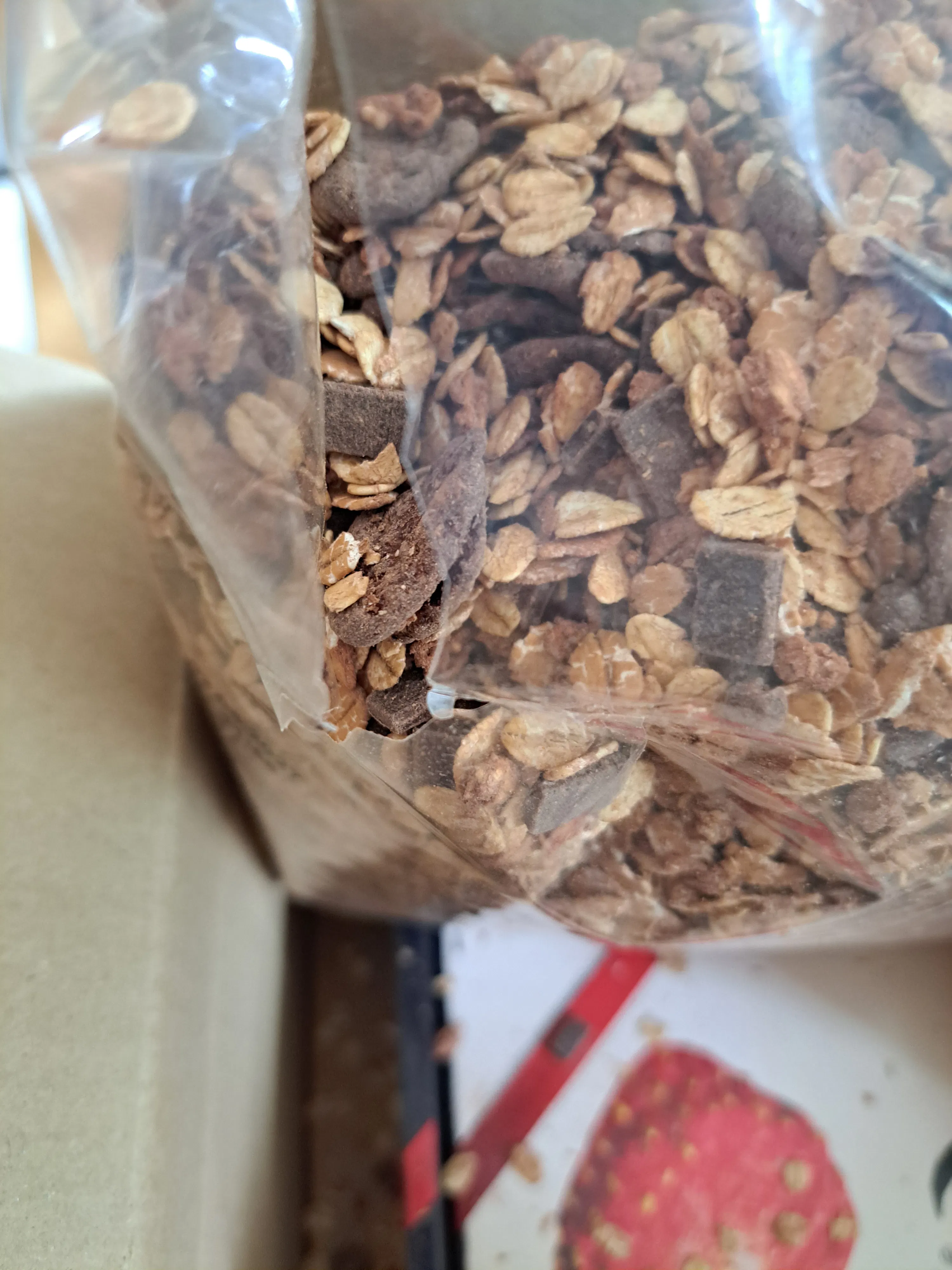 eine nahaufnahme einer durchsichtigen plastiktüte, gefüllt mit haferflocken-schokoladen-granola, mit einem unscharfen bild einer erdbeere auf einer verpackung im hintergrund.