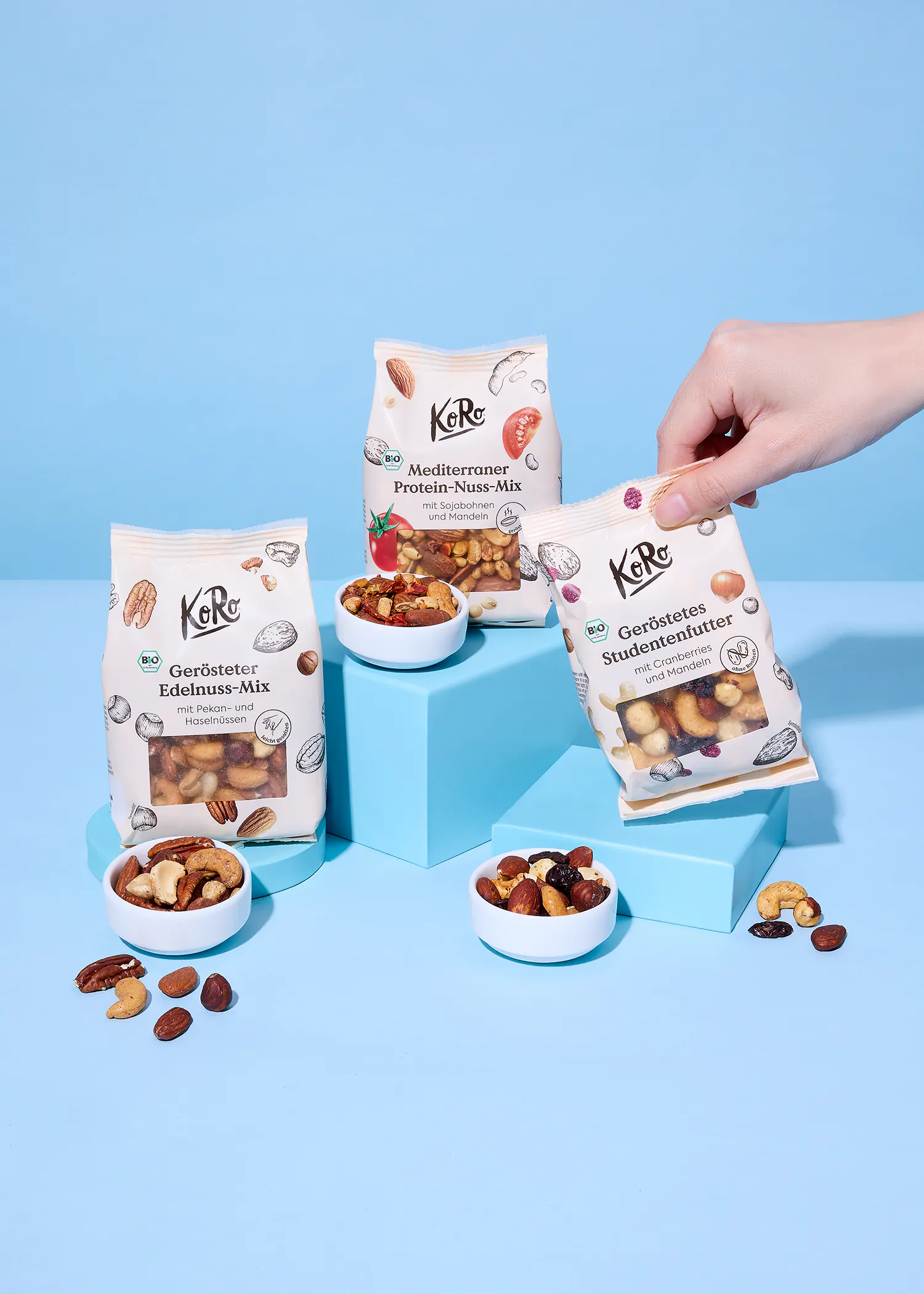 eine hand hält einen beutel koro geröstetes studentenfutter, während zwei weitere beutel (gerösteter edelnuss-mix und mediterraner protein-nuss-mix) auf blauen blöcken stehen, umgeben von kleinen schalen mit nussmischungen und verstreuten nüssen.