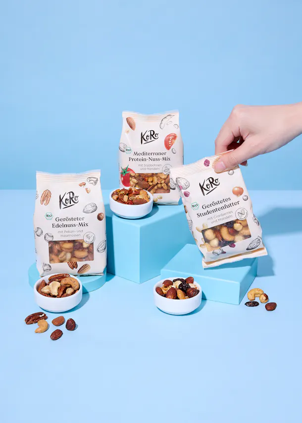 eine hand hält einen beutel koro geröstetes studentenfutter, während zwei weitere beutel (gerösteter edelnuss-mix und mediterraner protein-nuss-mix) auf blauen blöcken stehen, umgeben von kleinen schalen mit nussmischungen und verstreuten nüssen.