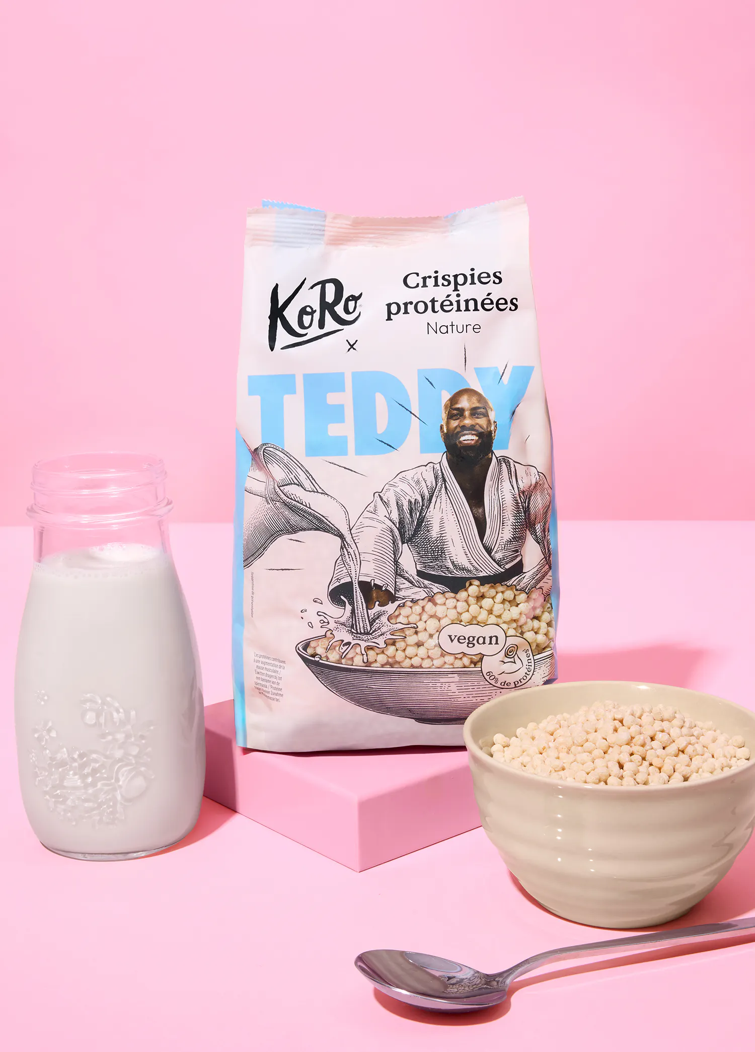 un sac de crispies protéinées koro nature avec une illustration de teddy riner versant du lait dans un bol de crispies, accompagné d'une bouteille de lait, d'un bol de crispies et d'une cuillère sur un fond rose.