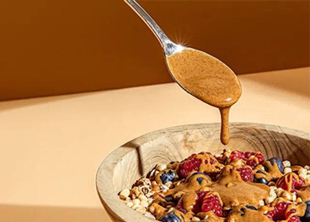 ein Löffel , der Erdnussbutter über eine Holzschale mit Müsli und Beeren gießt
