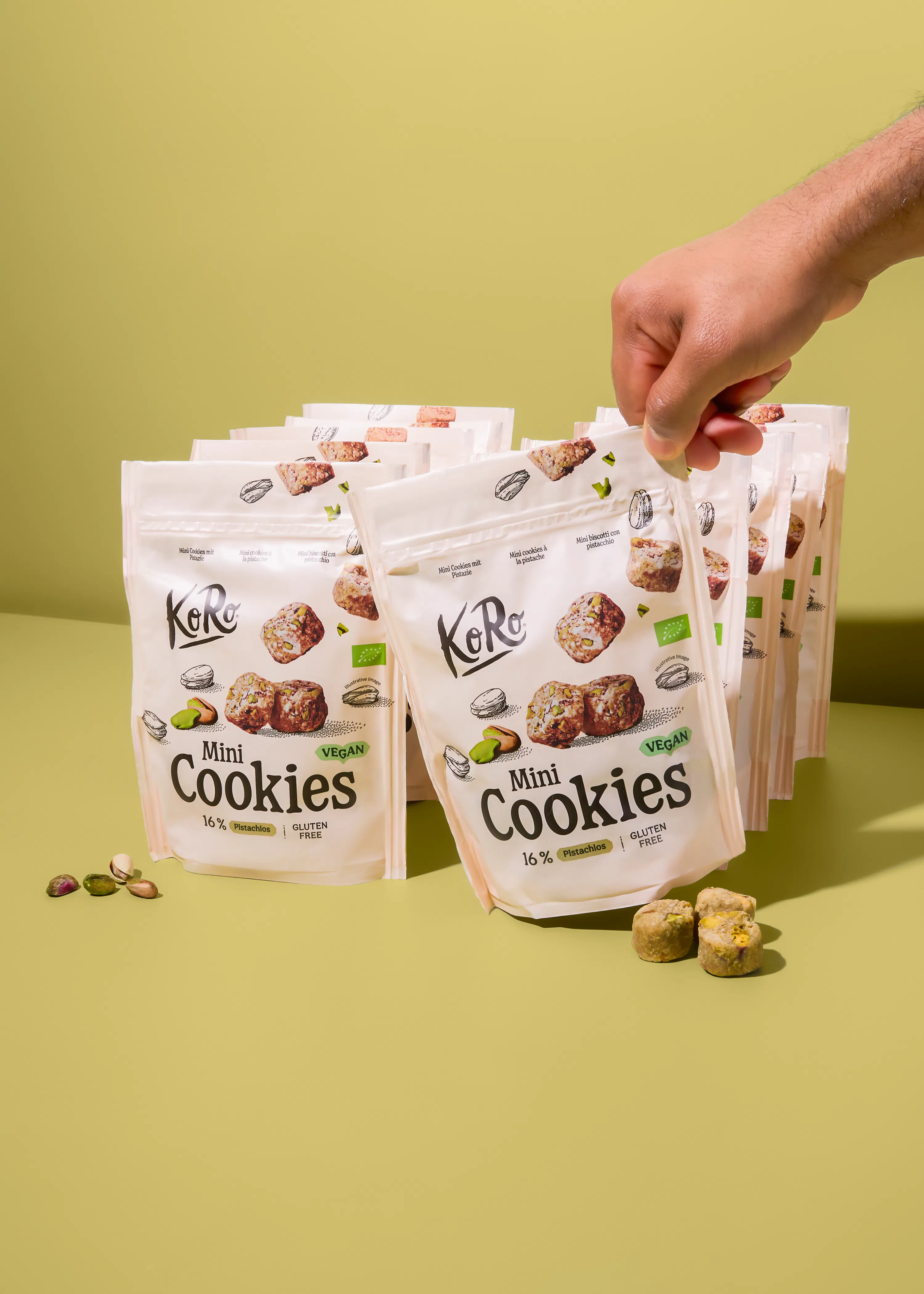 eine hand greift nach einer weißen packung koro mini cookies mit pistazien, vegan und glutenfrei, umgeben von weiteren packungen, losen pistazien und zwei keksen auf einem hellgrünen hintergrund.