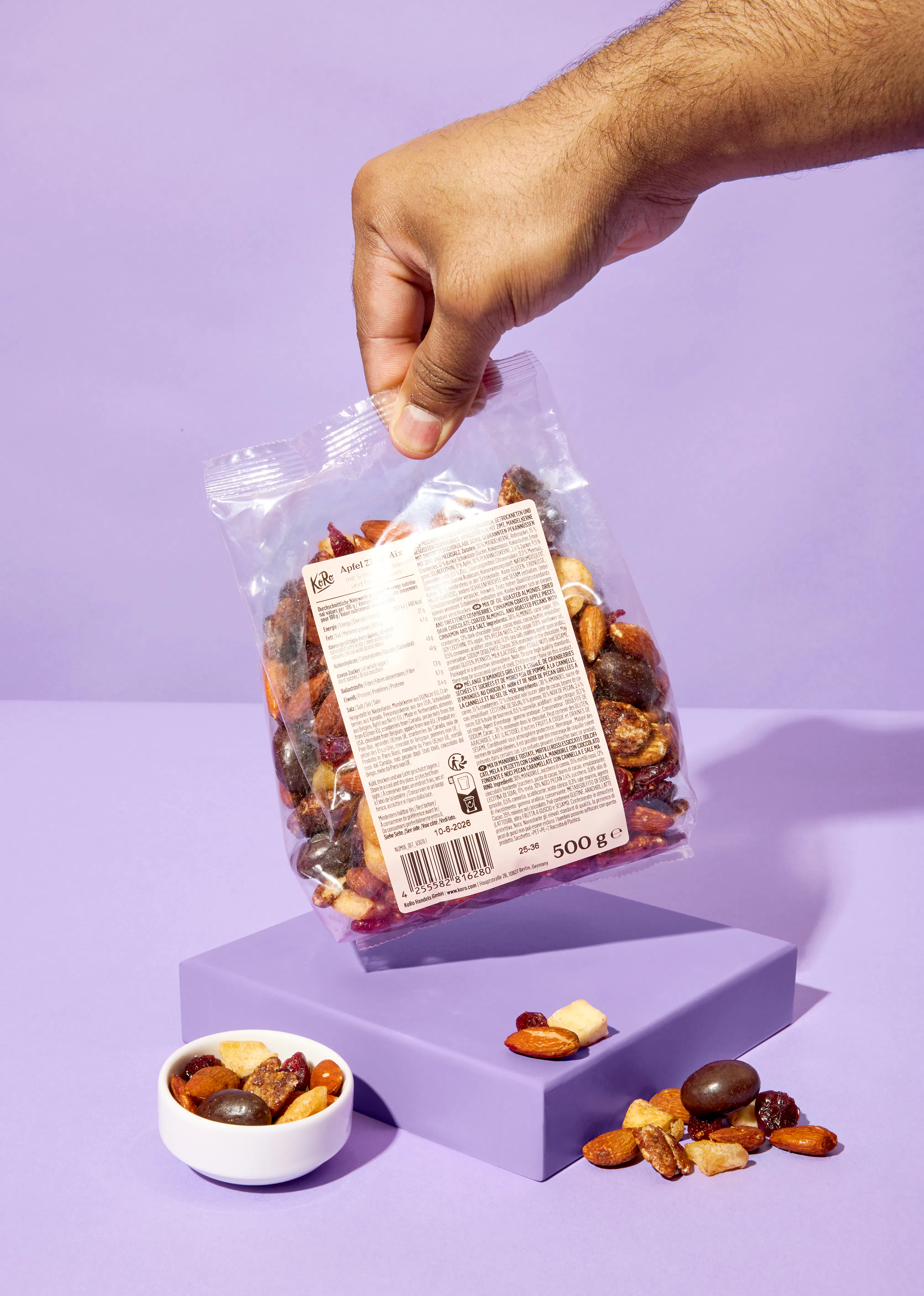 une main tient un sachet transparent de mélange de fruits secs et de noix au-dessus d'une surface violette, avec un bol et des morceaux éparpillés du mélange.