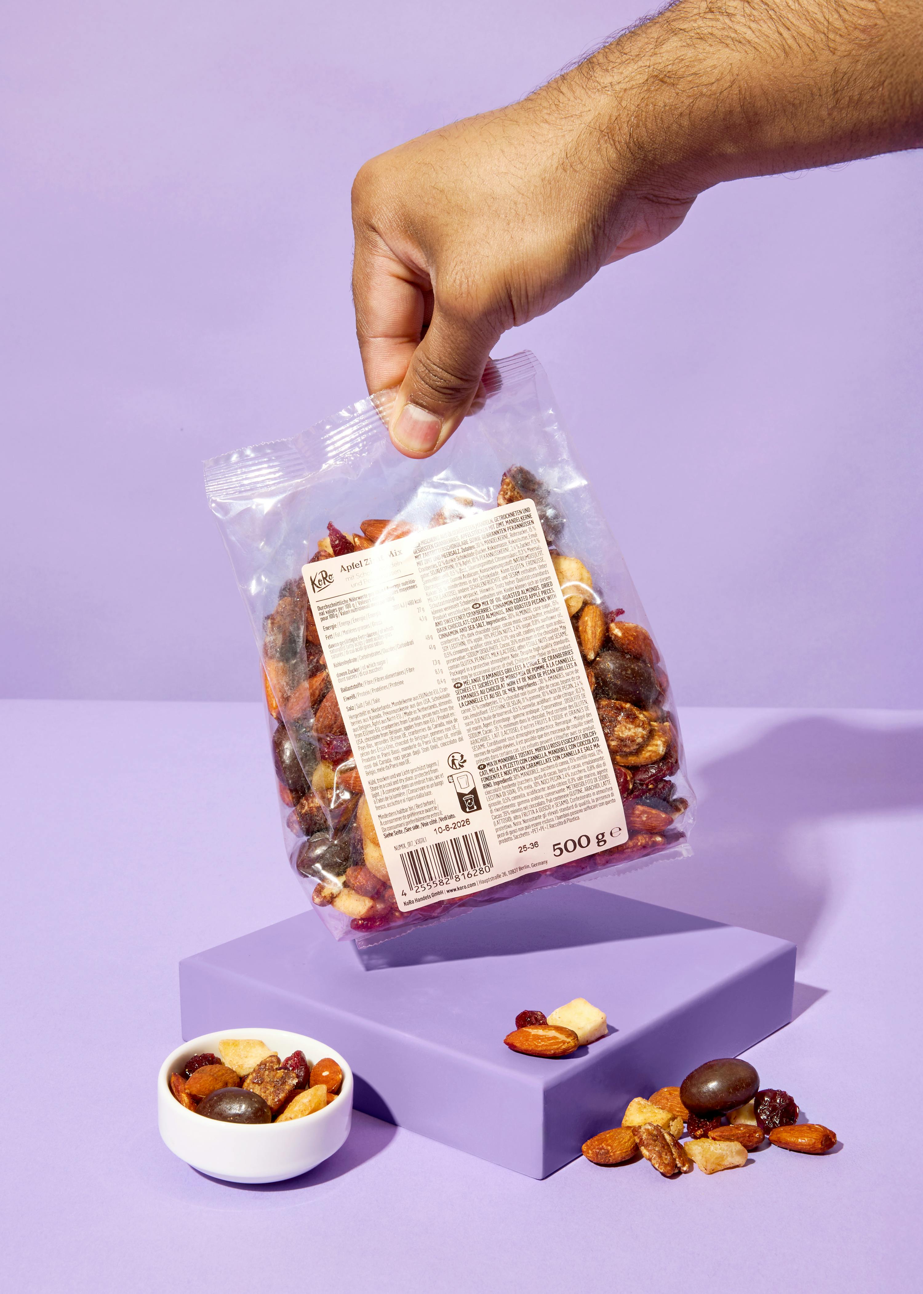a hand holds a clear bag of mixed nuts and dried fruit over a purple block, with some of the mix in a white bowl and spilled on the block.