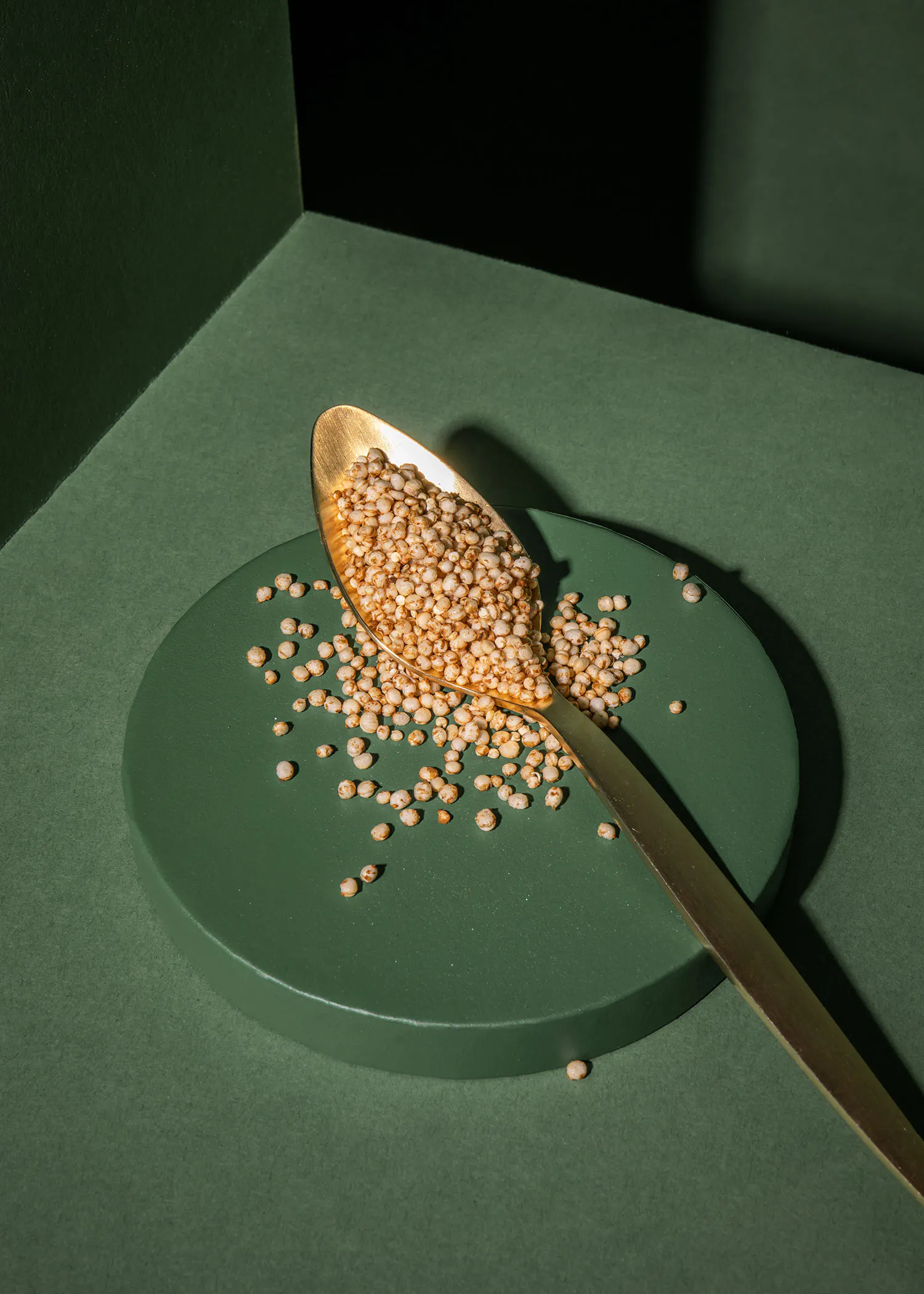 una cuchara dorada llena de granos de quinua inflada sobre un plato circular verde oscuro con algunos granos esparcidos y un fondo verde oscuro con sombras.