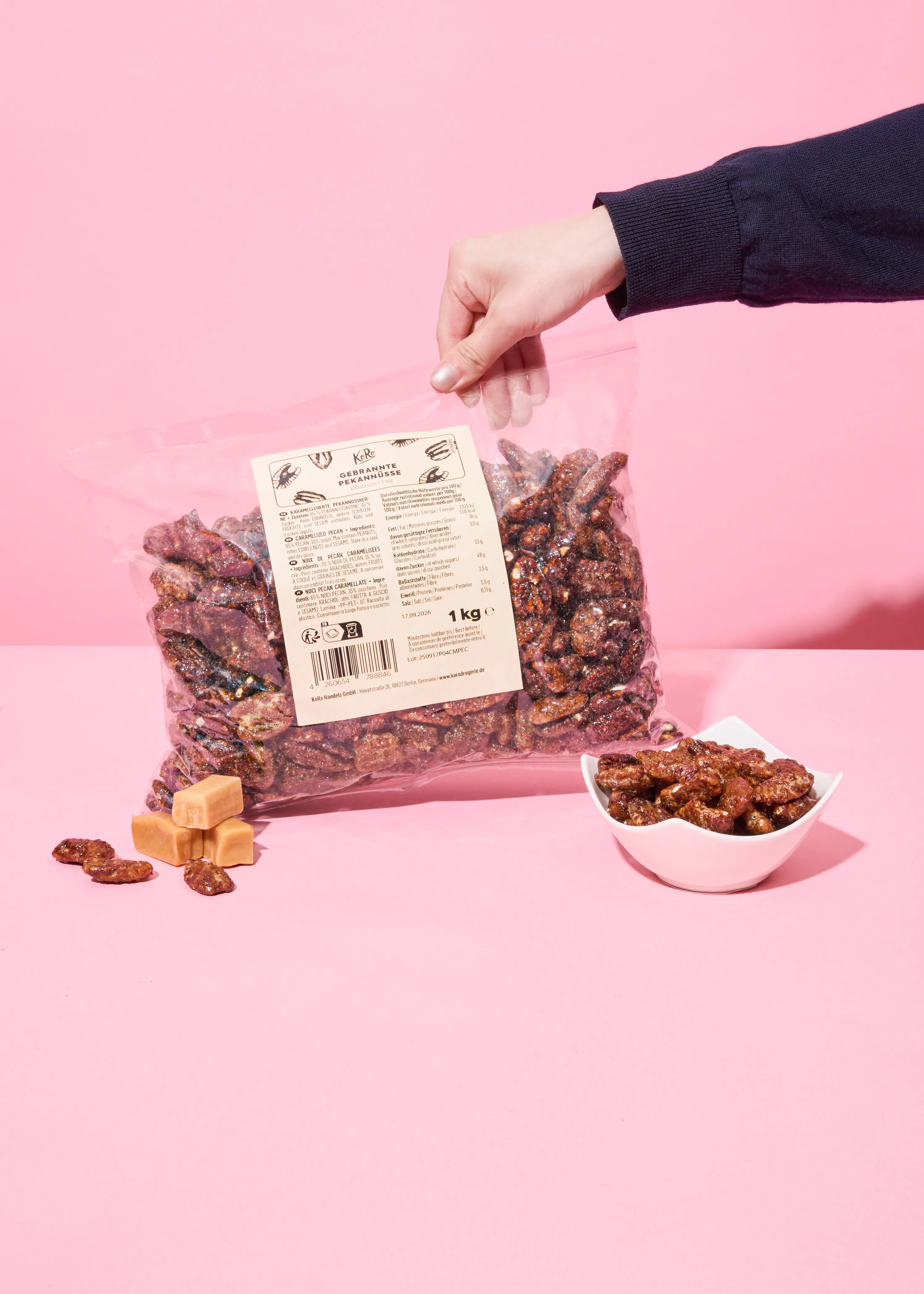 une main tient un grand sac transparent de 1 kg de noix de pécan caramélisées, avec un petit bol de noix de pécan et des bonbons au caramel posés sur une surface rose.