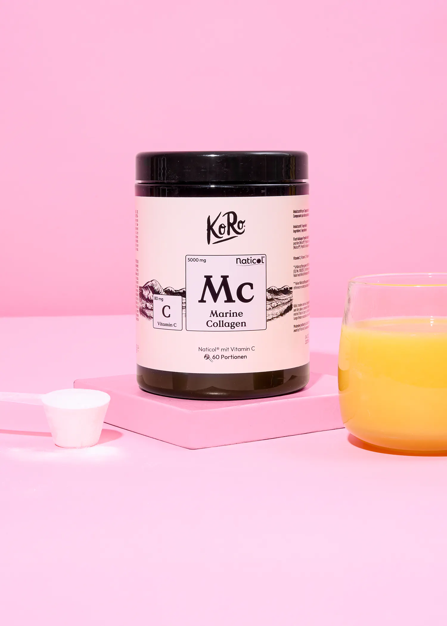 a black and white container of koro naticol marine collagen with vitamin c, a white scoop of powder, and a glass of orange juice are displayed on a pink stand against a pink background.