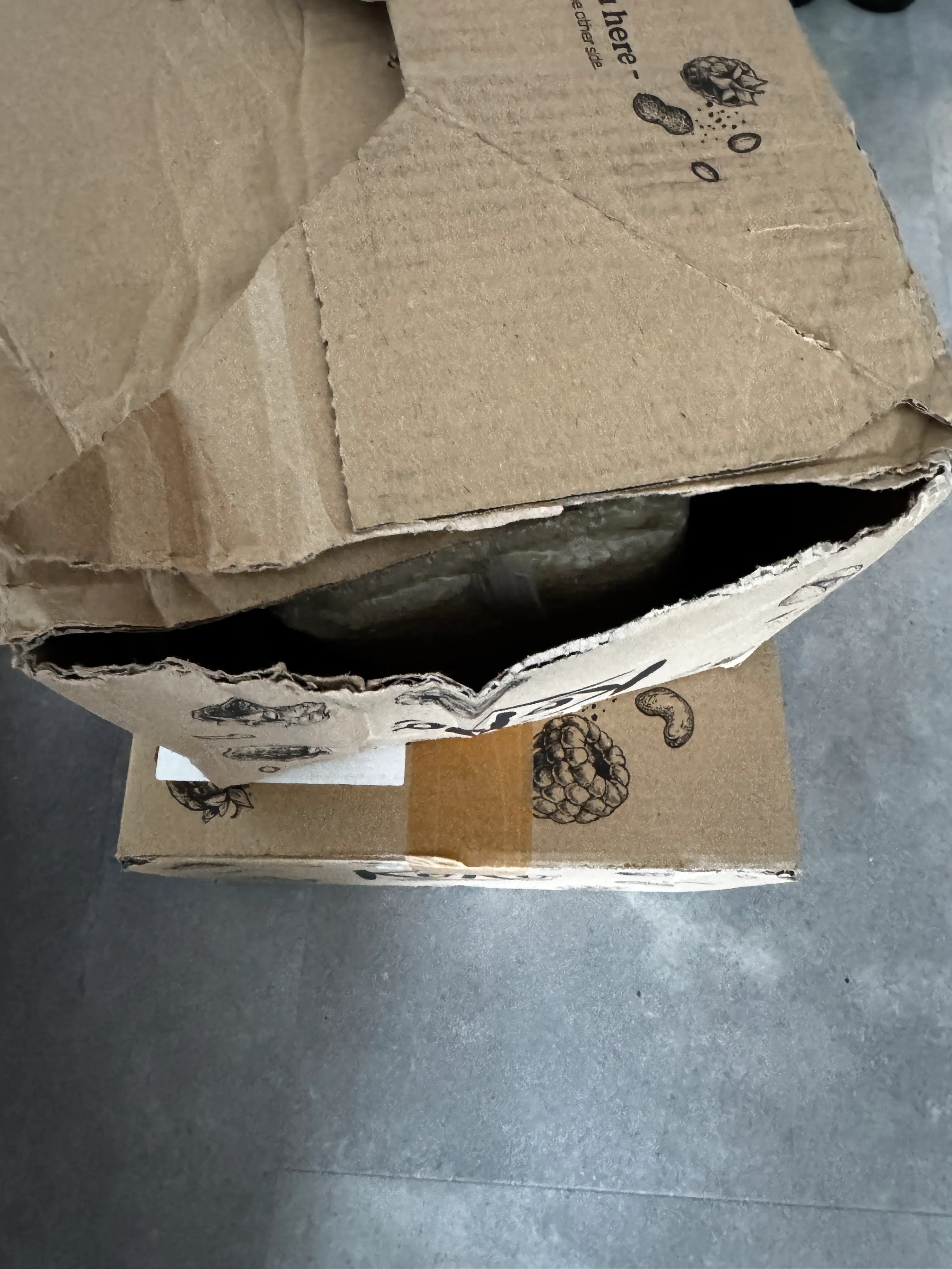 zwei braune kartons, von denen der vordere oben offen und eingerissen ist und beide mit schwarzen illustrationen von nüssen und beeren bedruckt sind, stehen auf einem grauen boden.