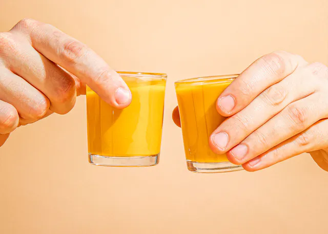 zwei hände halten zwei kleine gläser mit gelbem saft