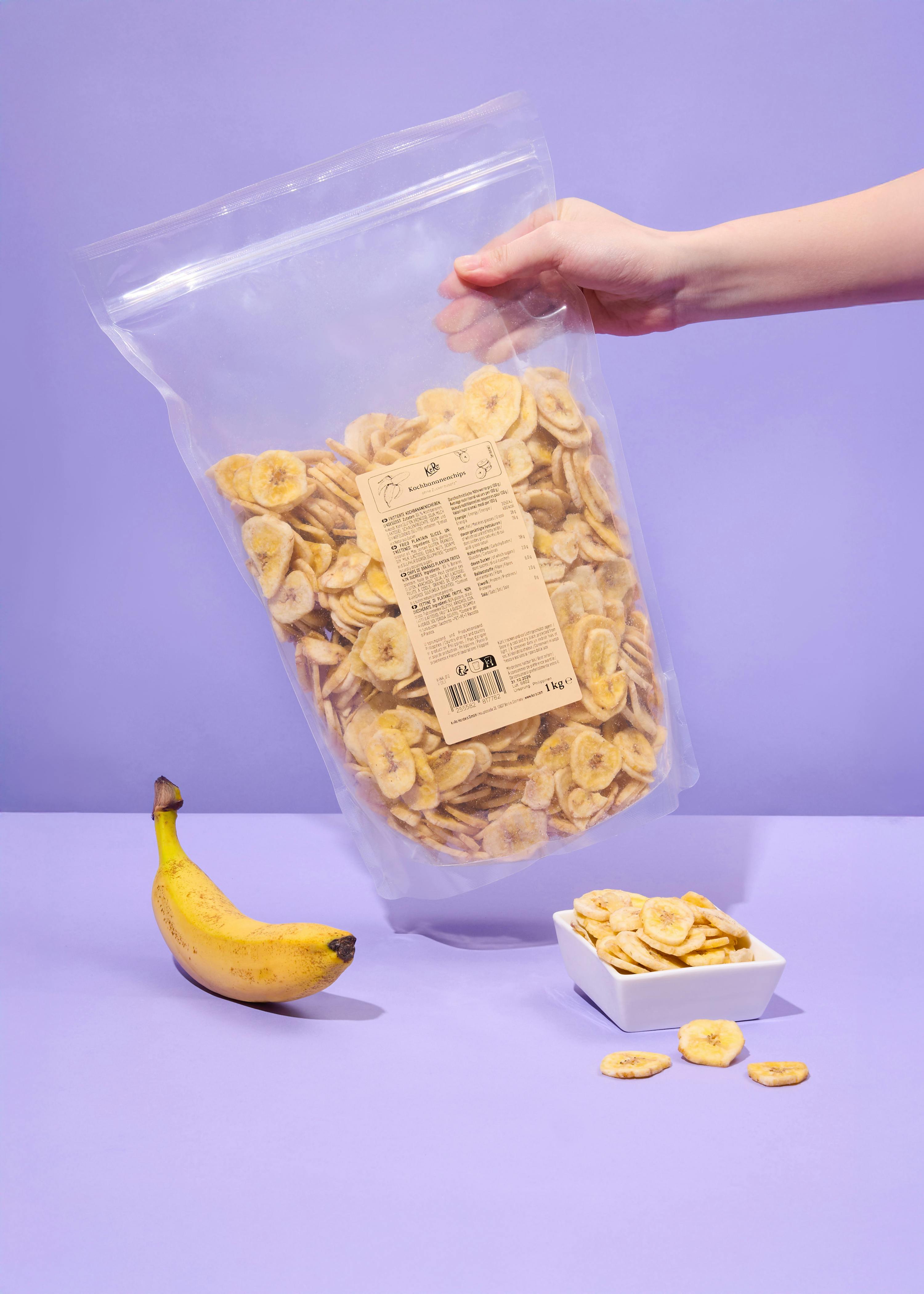 una mano tiene una grande busta trasparente di kochbananenchips, con una banana intera e una ciotola di chips su uno sfondo viola.