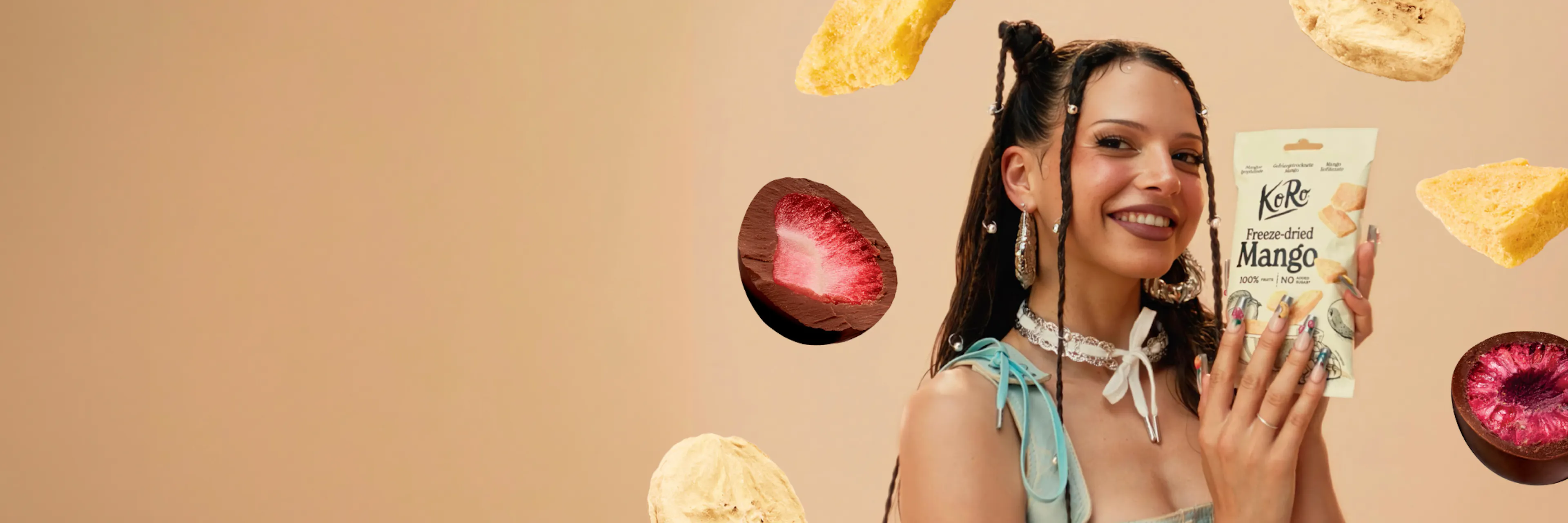 eine junge frau mit geflochtenen haaren hält lächelnd eine packung gefriergetrocknete mango von koro in die kamera, umgeben von schwebenden stücken gefriergetrockneter mango und schokolierten früchten vor einem beigen hintergrund.