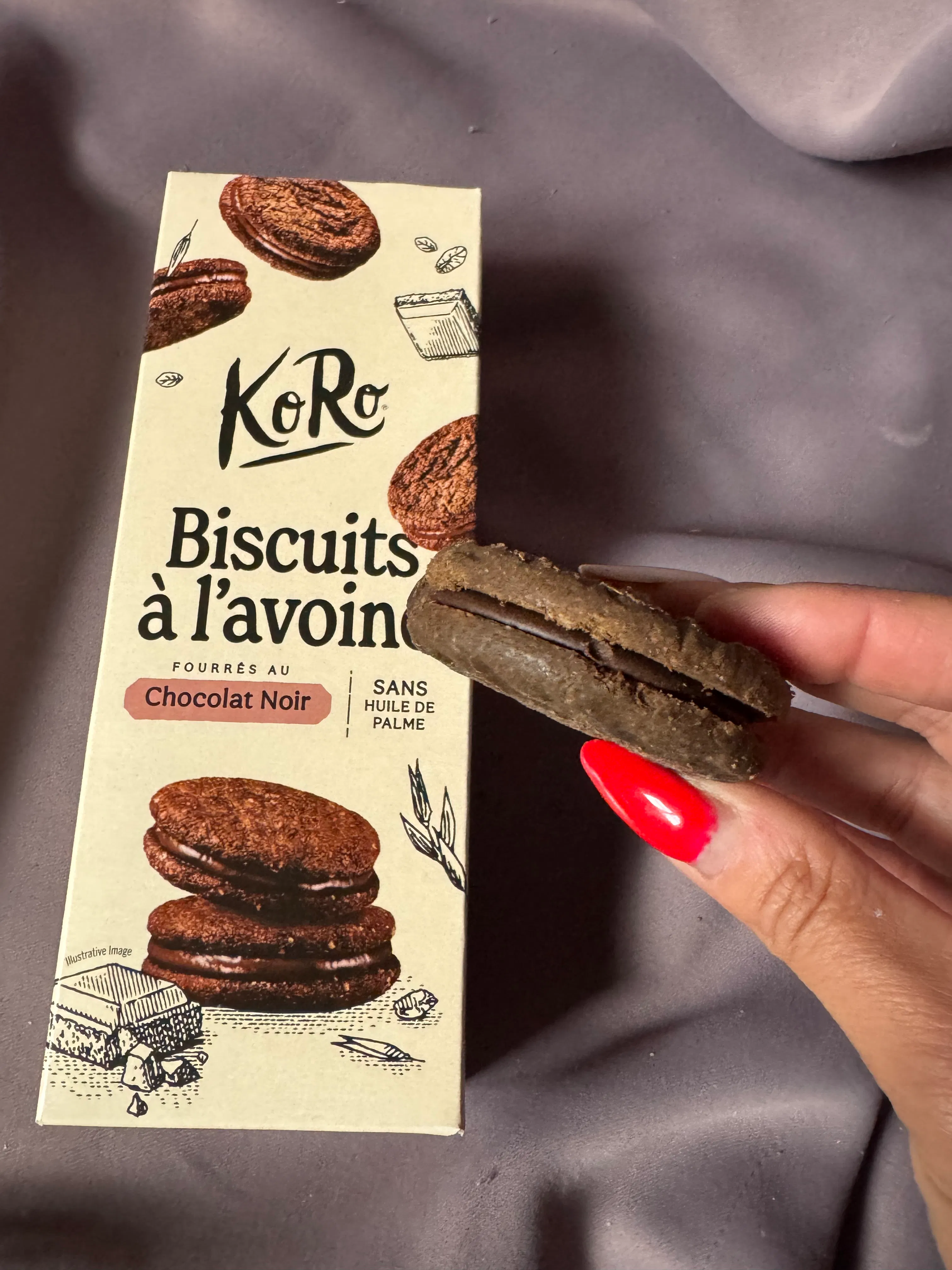 una mano con unghie rosse tiene un biscotto all'avena farcito al cioccolato sopra la sua confezione koro.