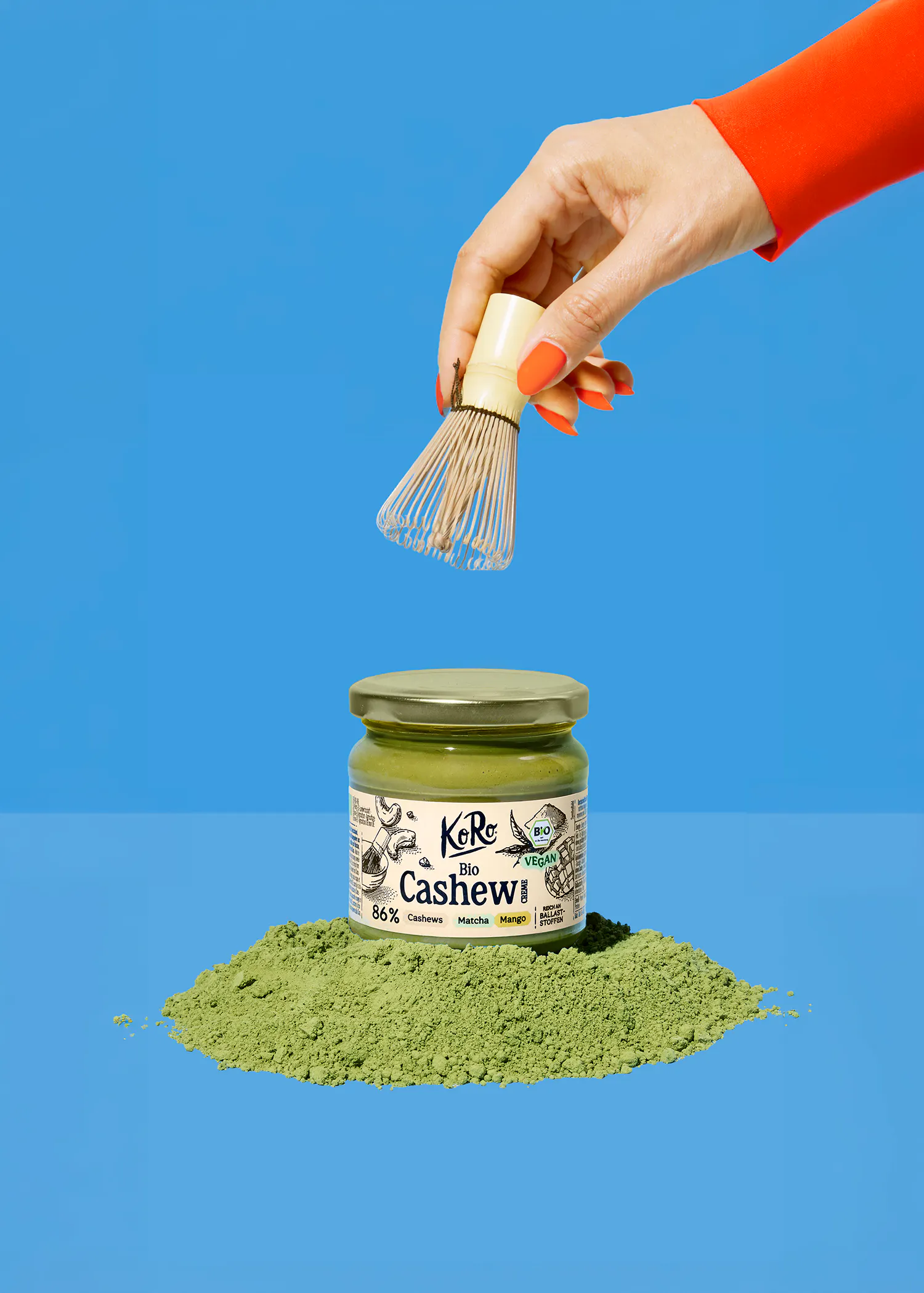 eine hand mit orangefarbenen nägeln hält einen matcha-besen über einem glas koro bio cashew-aufstrich, der auf einem haufen grünem pulver auf blauem hintergrund steht.