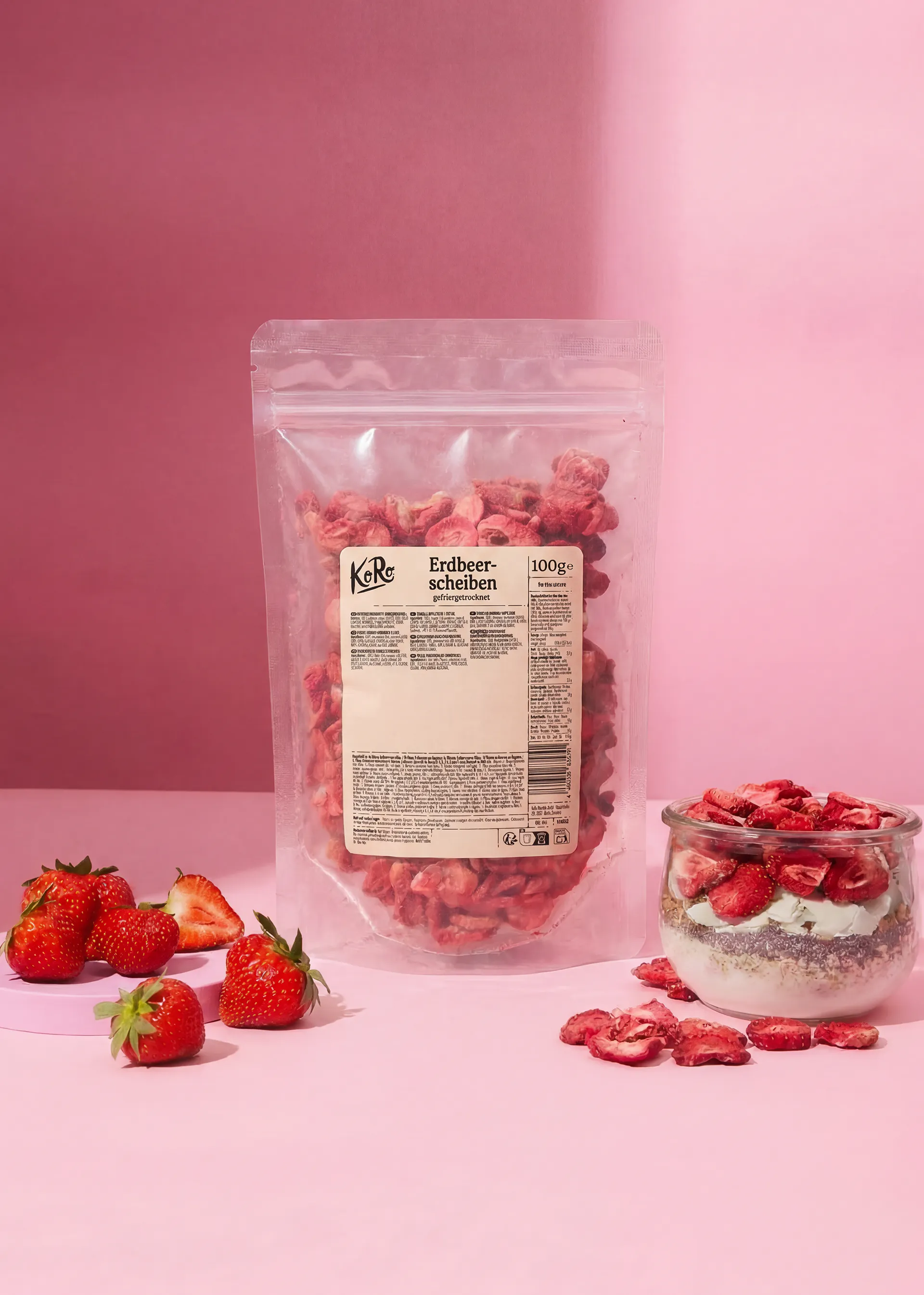 eine transparente verpackung mit gefriergetrockneten erdbeerscheiben, frische erdbeeren und ein glas mit joghurt und erdbeeren auf einem pinken hintergrund.