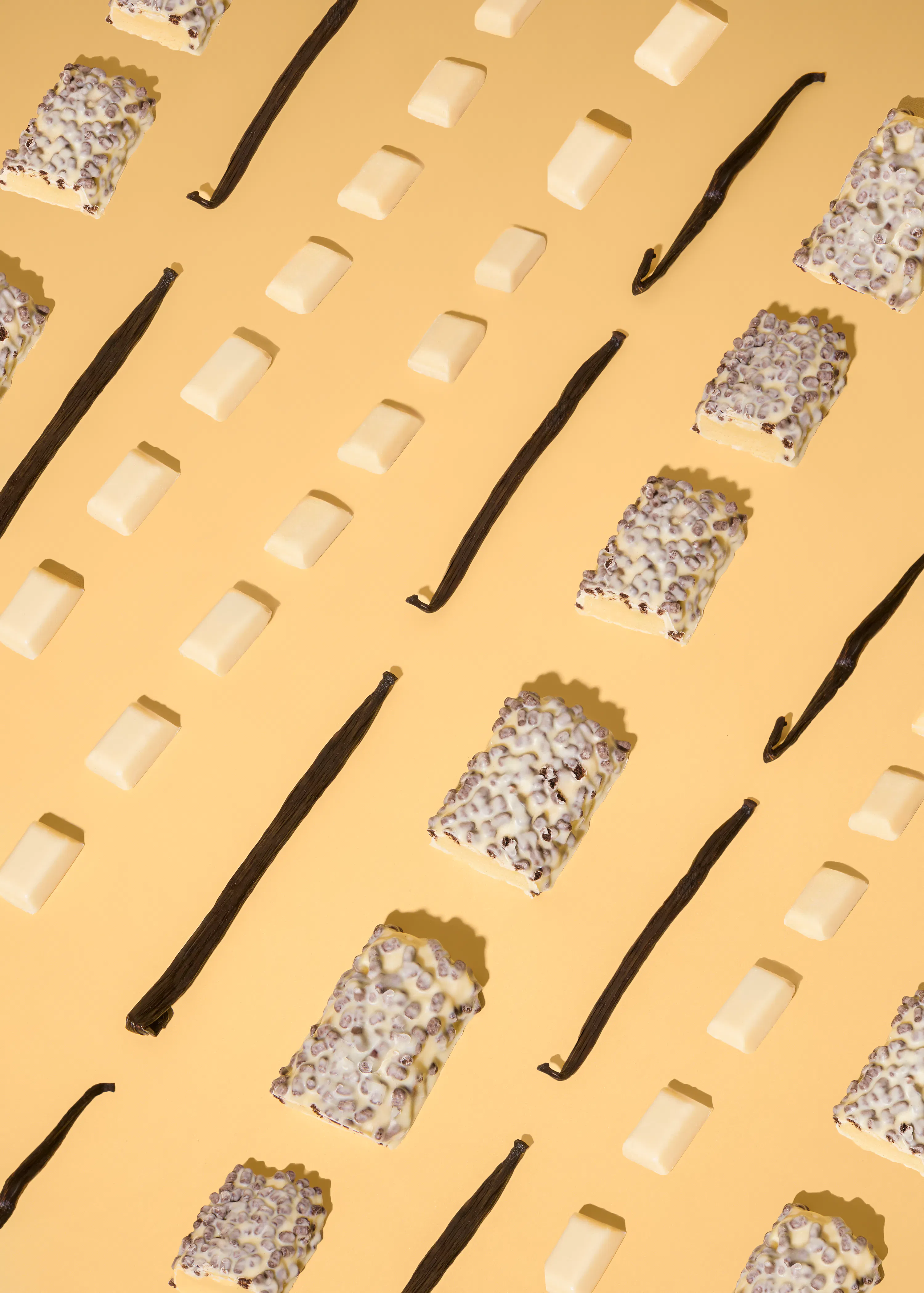 un motivo ripetuto di barrette di cioccolato bianco, baccelli di vaniglia e barrette di cioccolato bianco con scaglie di cioccolato su uno sfondo giallo chiaro.