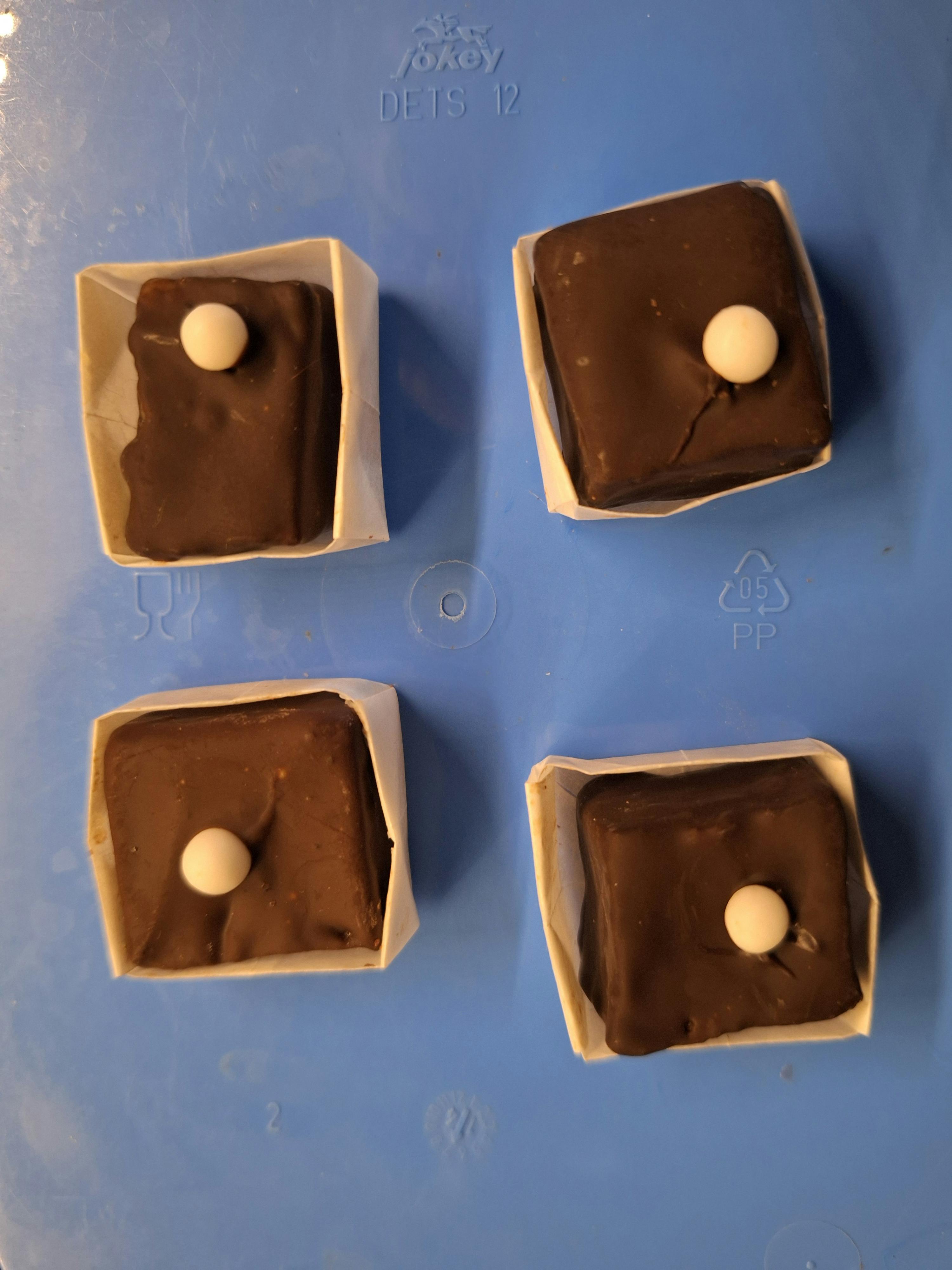 quatre chocolats carrés foncés, chacun dans une coupelle en papier blanc et surmonté d'une petite perle blanche, posés sur une surface bleue.