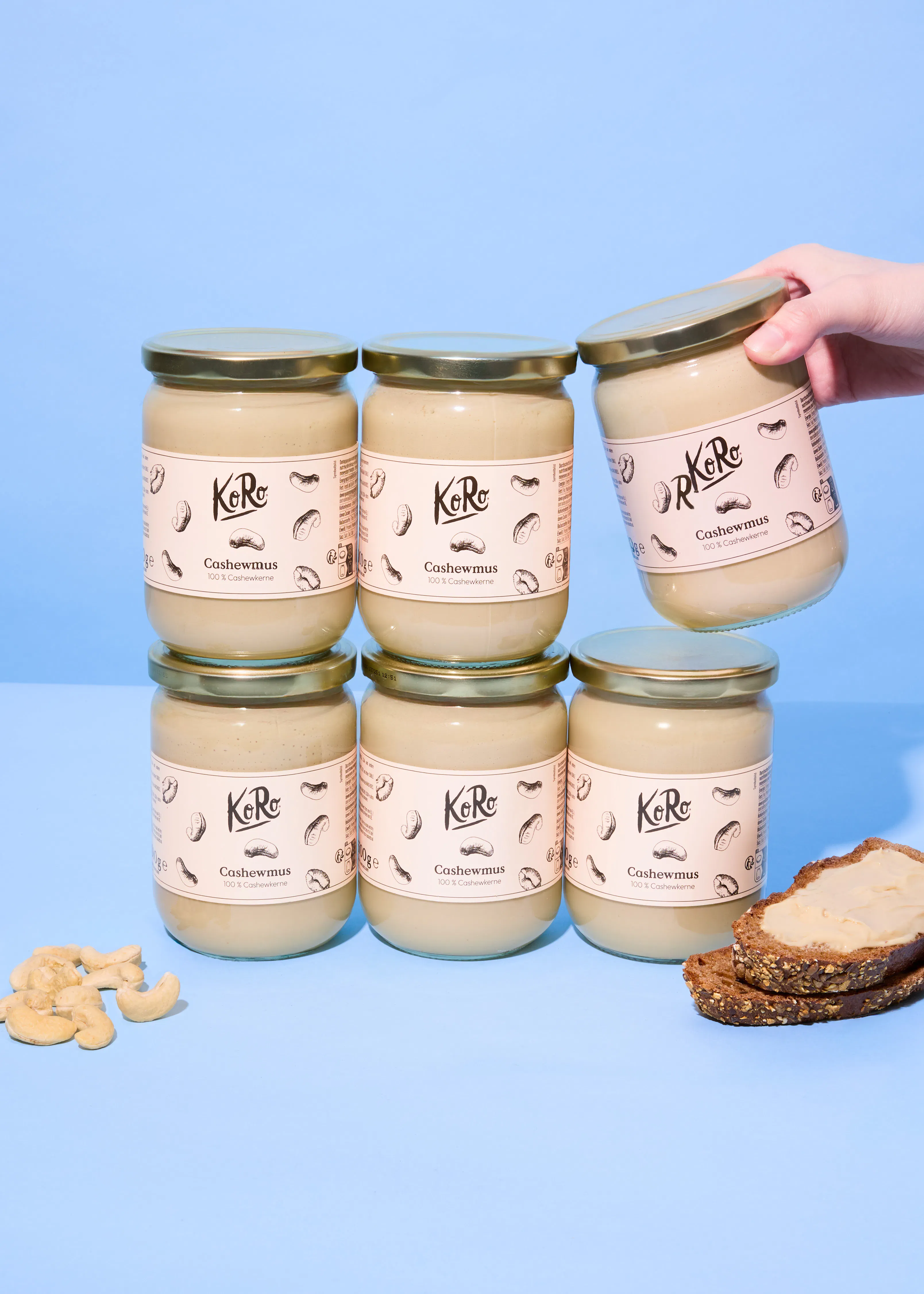 sechs gläser koro cashewmus, eines davon wird von einer hand gehalten, mit losen cashews und einem brot mit cashewmus auf blauem hintergrund.