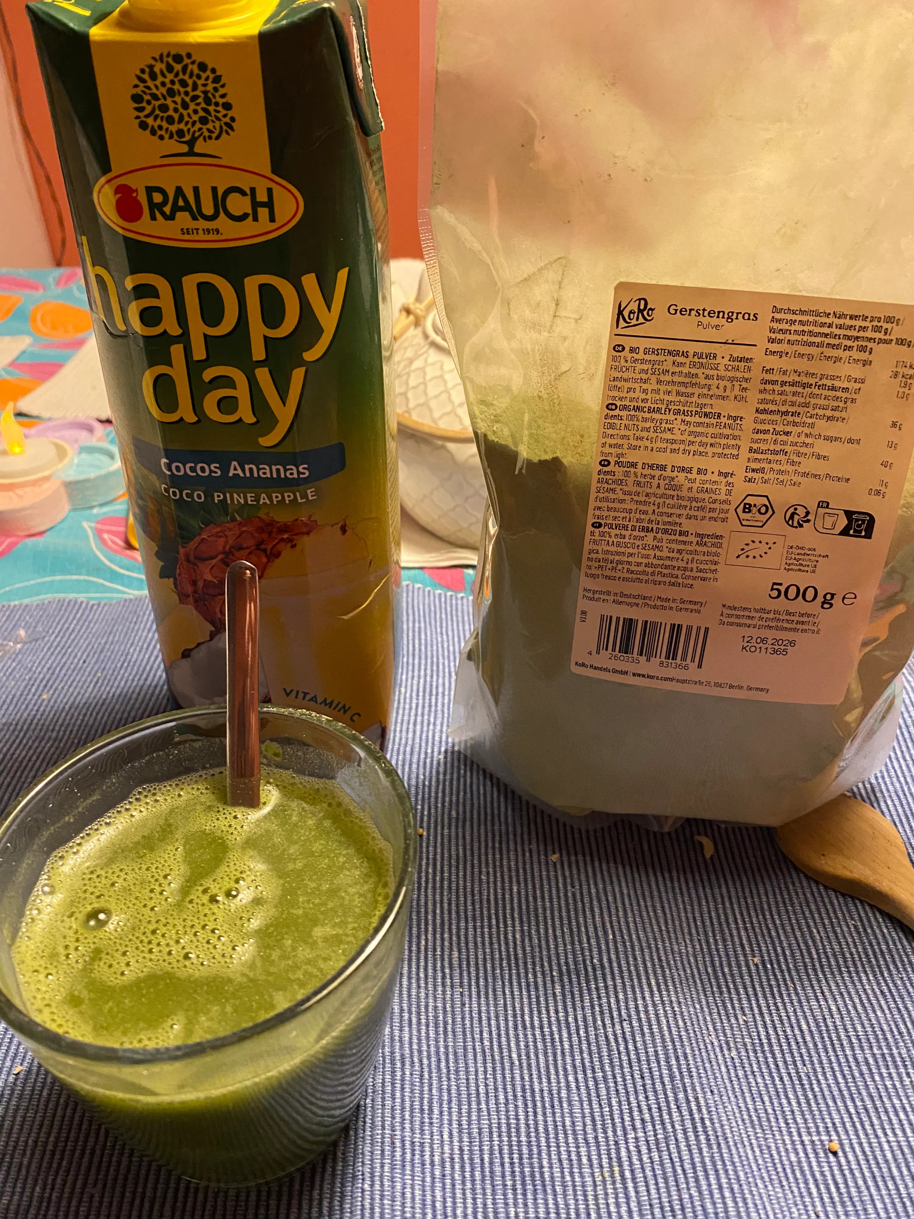ein glas mit grünem smoothie und strohhalm, daneben ein karton ananassaft und eine packung gerstengraspulver auf einem blau-weiß gestreiften tisch.