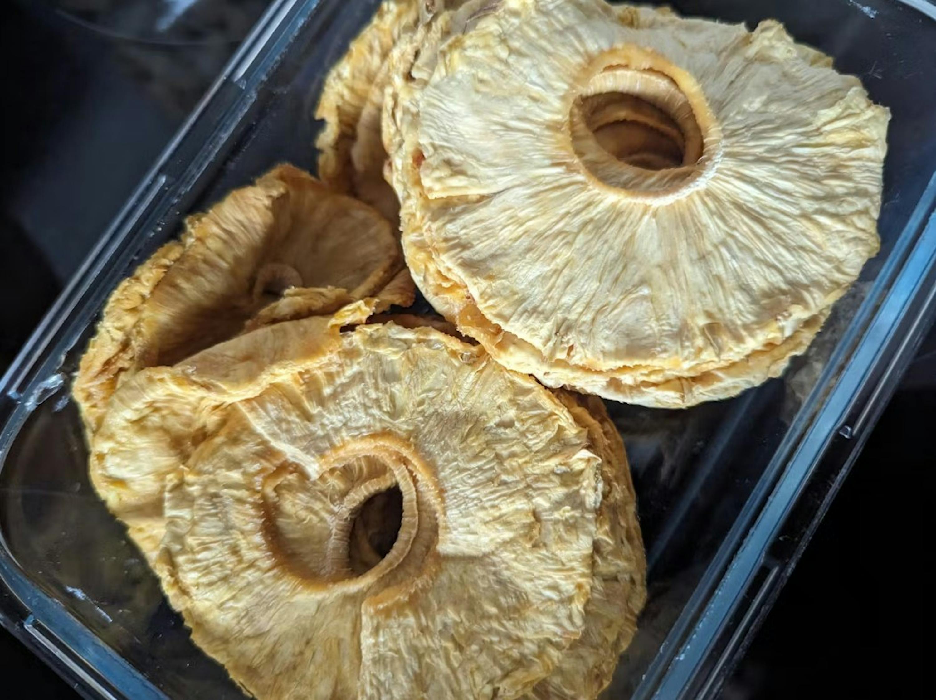diversi anelli di ananas essiccato di colore giallo chiaro con una superficie testurizzata sono impilati in un contenitore di plastica trasparente.