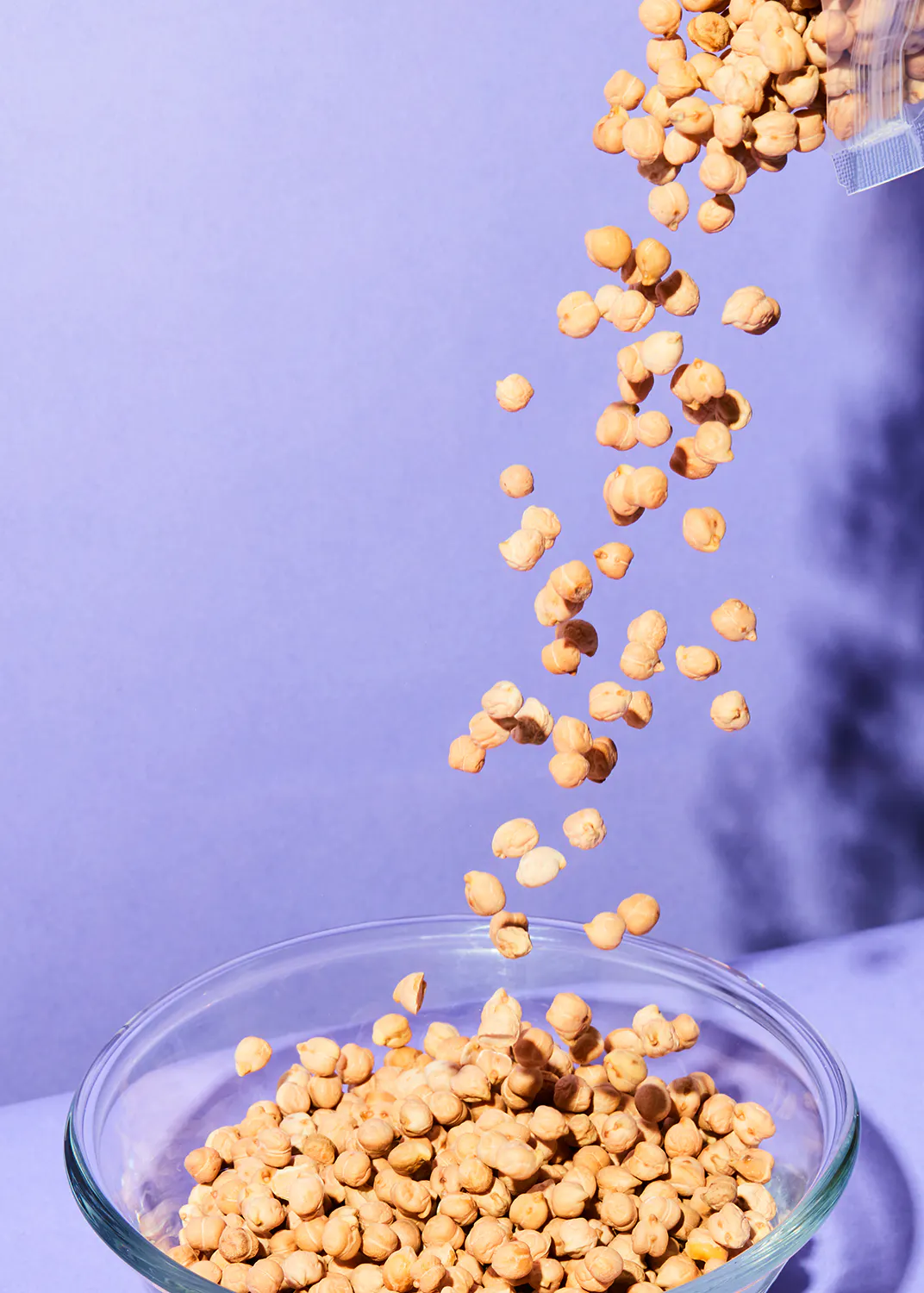 chickpeas being poured into a glass bowl on a purple background