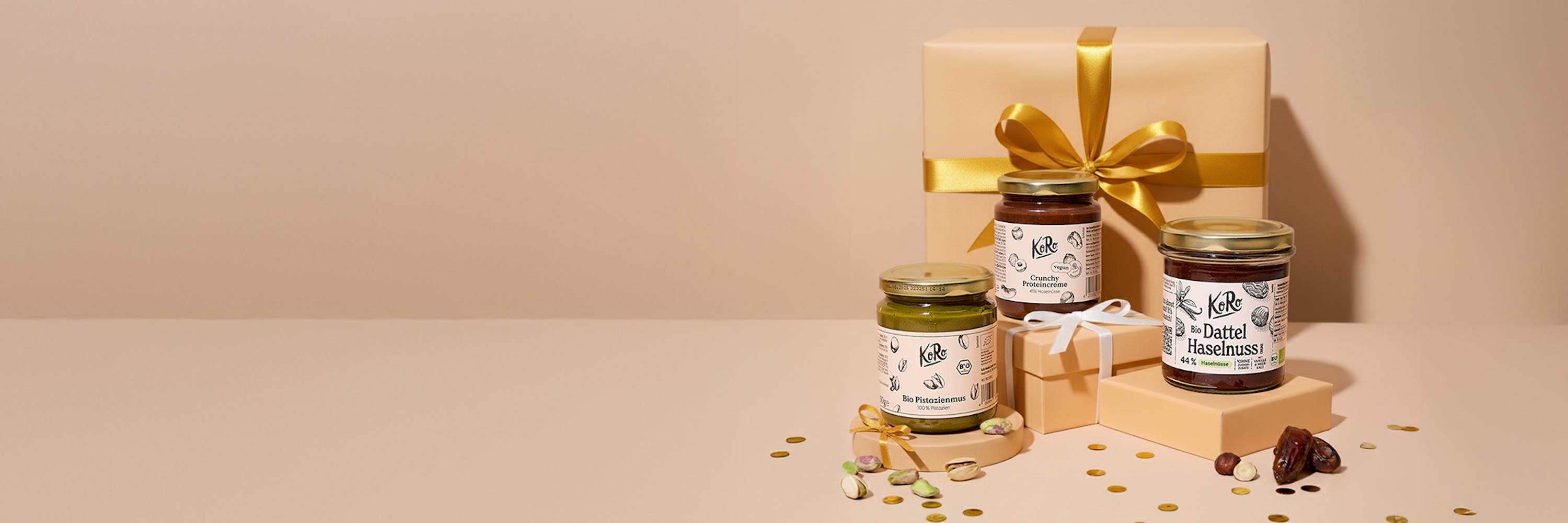 three koro brand jars of pistachio, crunchy protein, and date hazelnut spreads are displayed with gift boxes, nuts, and confetti on a light pink surface.