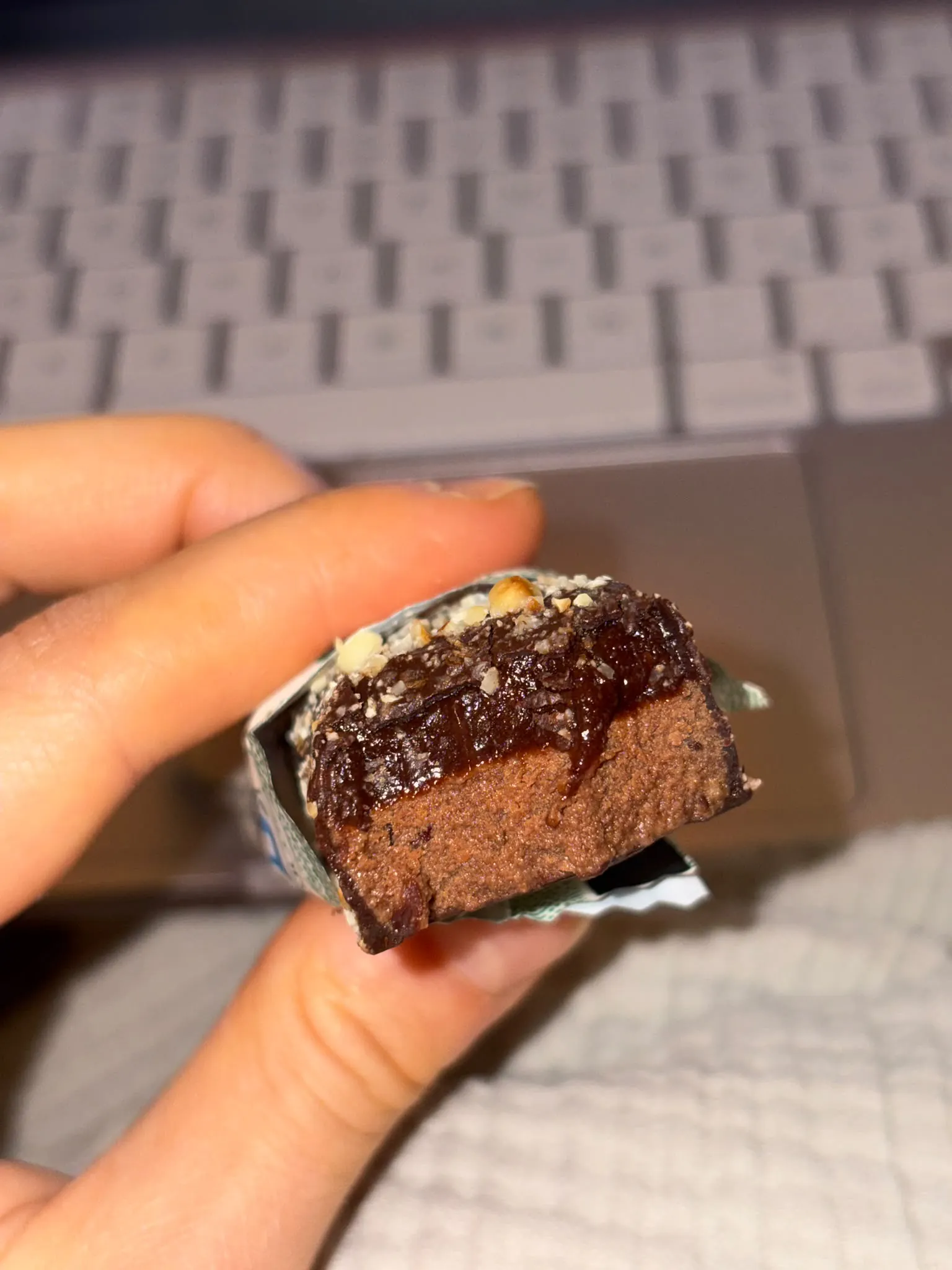 eine hand hält einen angebissenen schokoriegel mit einer schicht aus dunkler creme und gehackten nüssen vor einer laptop-tastatur.